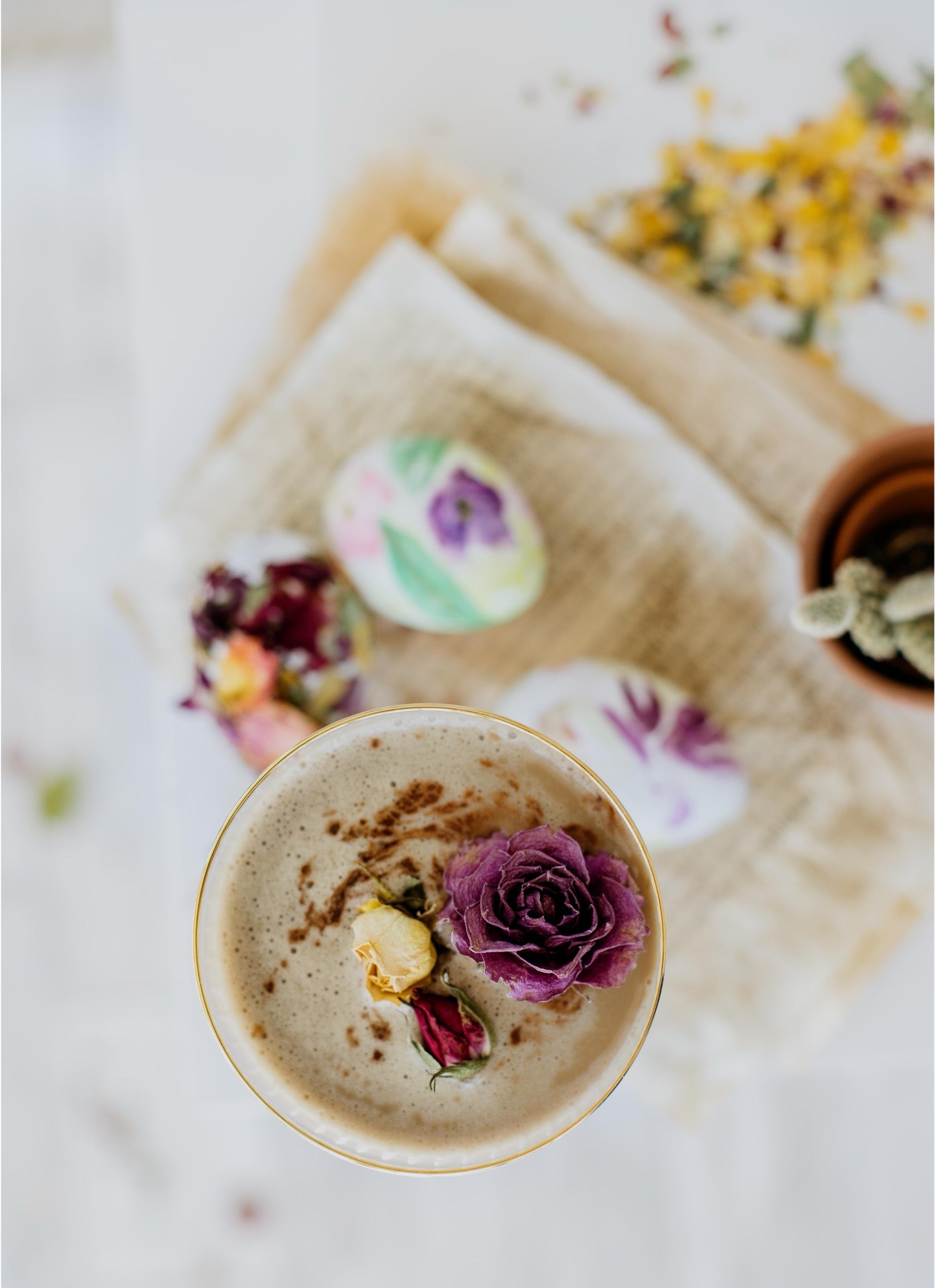 A top-down view of a frothy beige beverage in a clear glass with a gold rim, garnished with a purple rose, a small yellow flower, and a sprinkle of brown powder. The drink sits on a beige cloth with blurred decorative eggs and dried flowers in the background.
