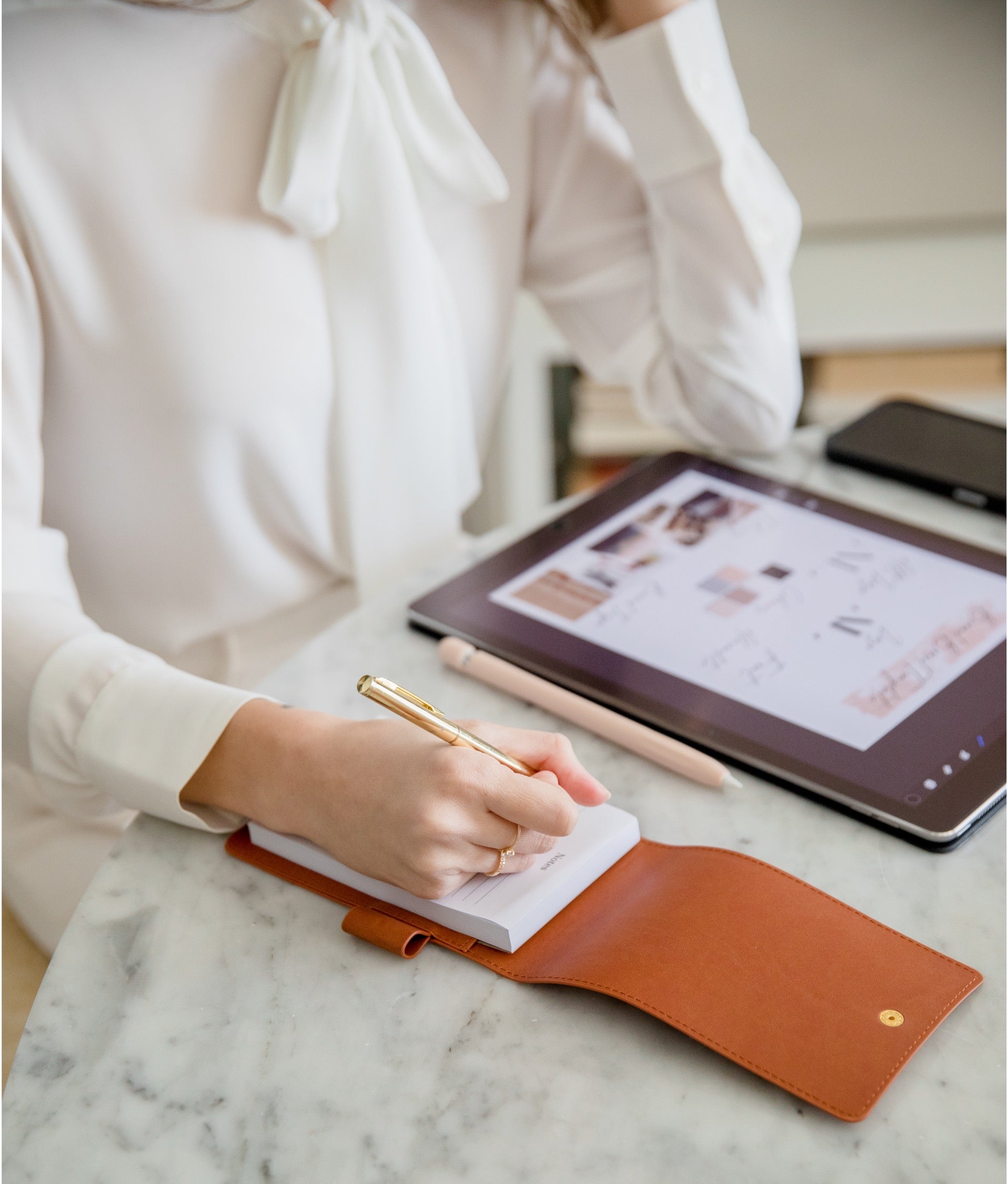 A person in a white blouse writes in a small notebook with a gold pen inside a brown leather cover on a marble desk, with a tablet and a pink stylus nearby.