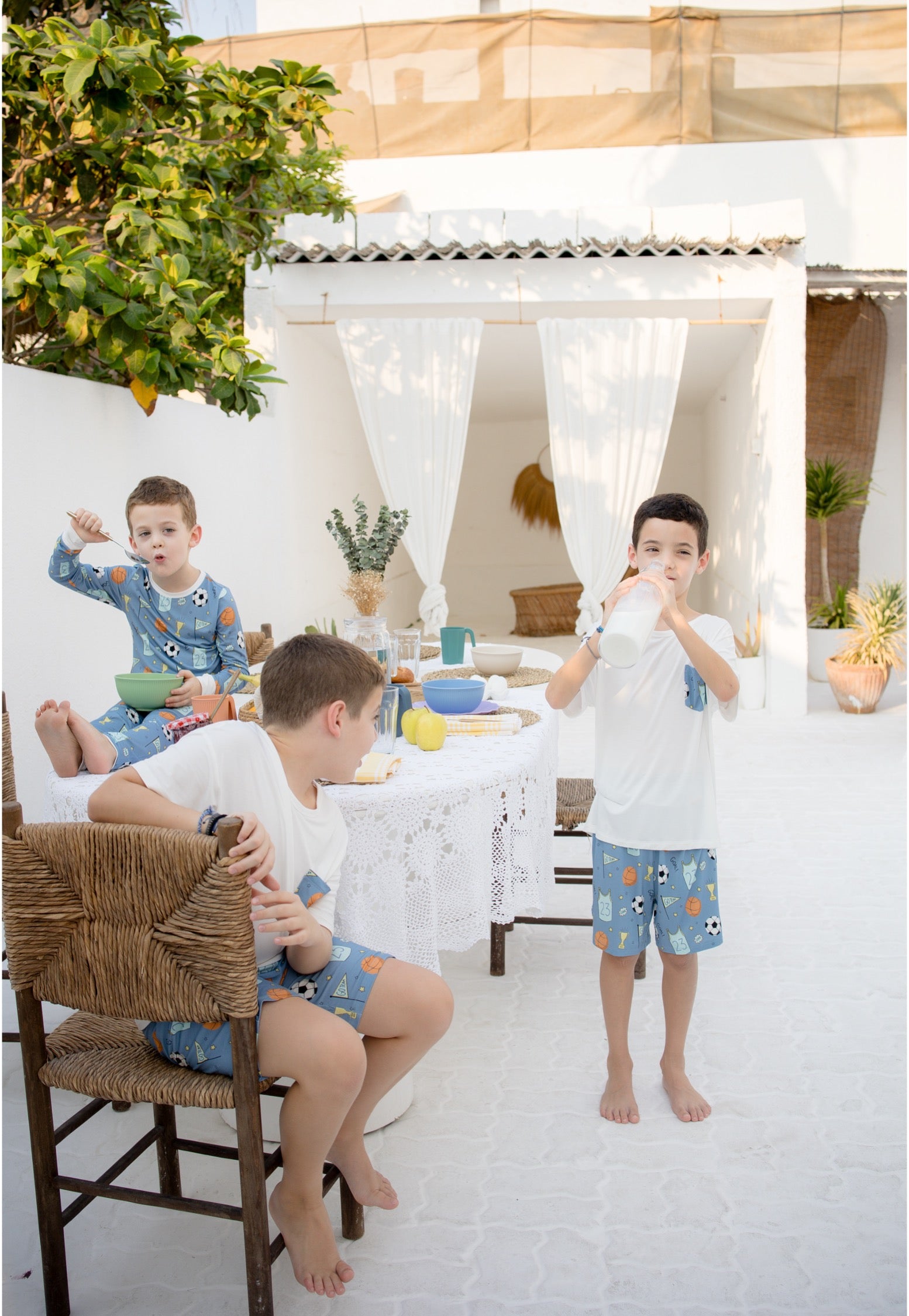Three children wearing matching blue patterned pajamas and shorts lounge in a bright, sunlit Mediterranean-style patio setting with white walls, a wicker chair, and a white pergola.