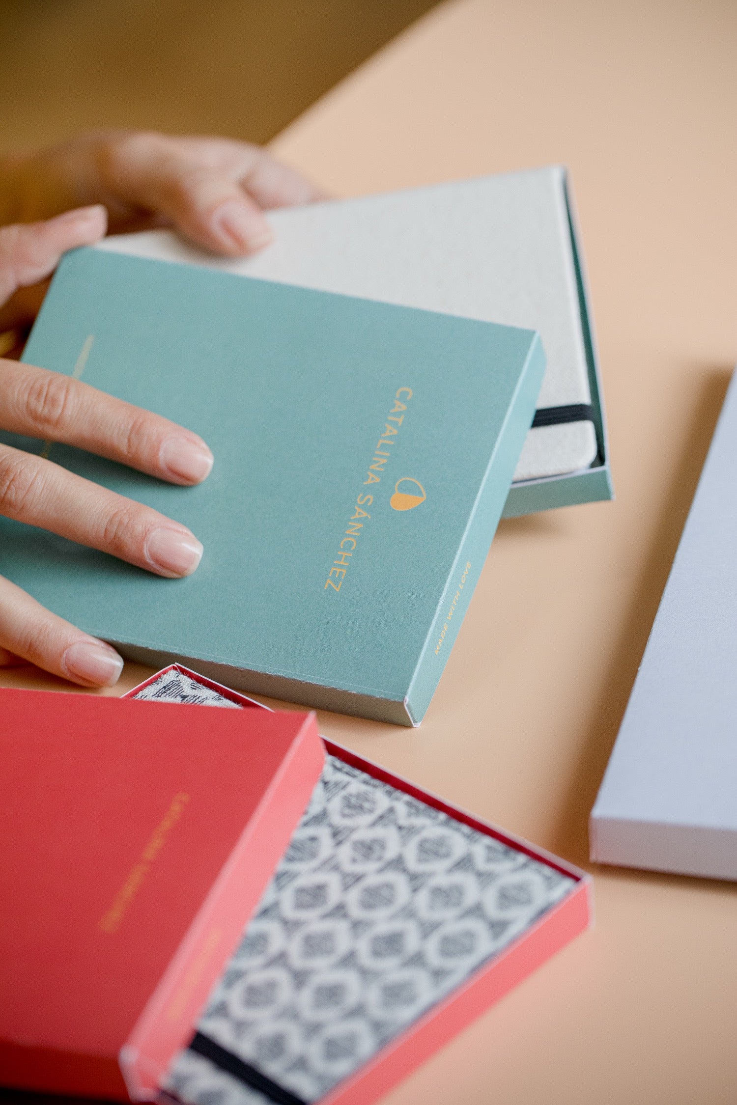 A close-up of hands opening a teal gift box with gold lettering, alongside other gift packaging in red and white on a warm beige surface.