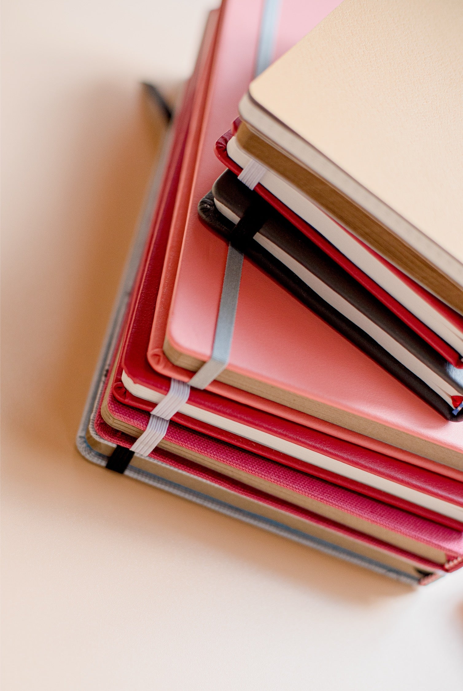 A stack of hardcover notebooks with colorful covers, including pink, red, black, beige, and gray, arranged diagonally on a light cream background.