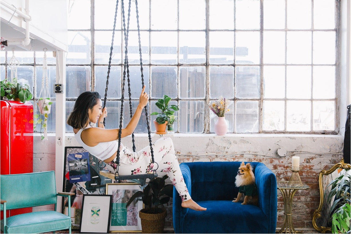 A woman wearing a white crop top and white floral print leggings sits on a hanging swing chair in a bright loft-style room with a blue velvet armchair, a small dog, and large industrial windows.