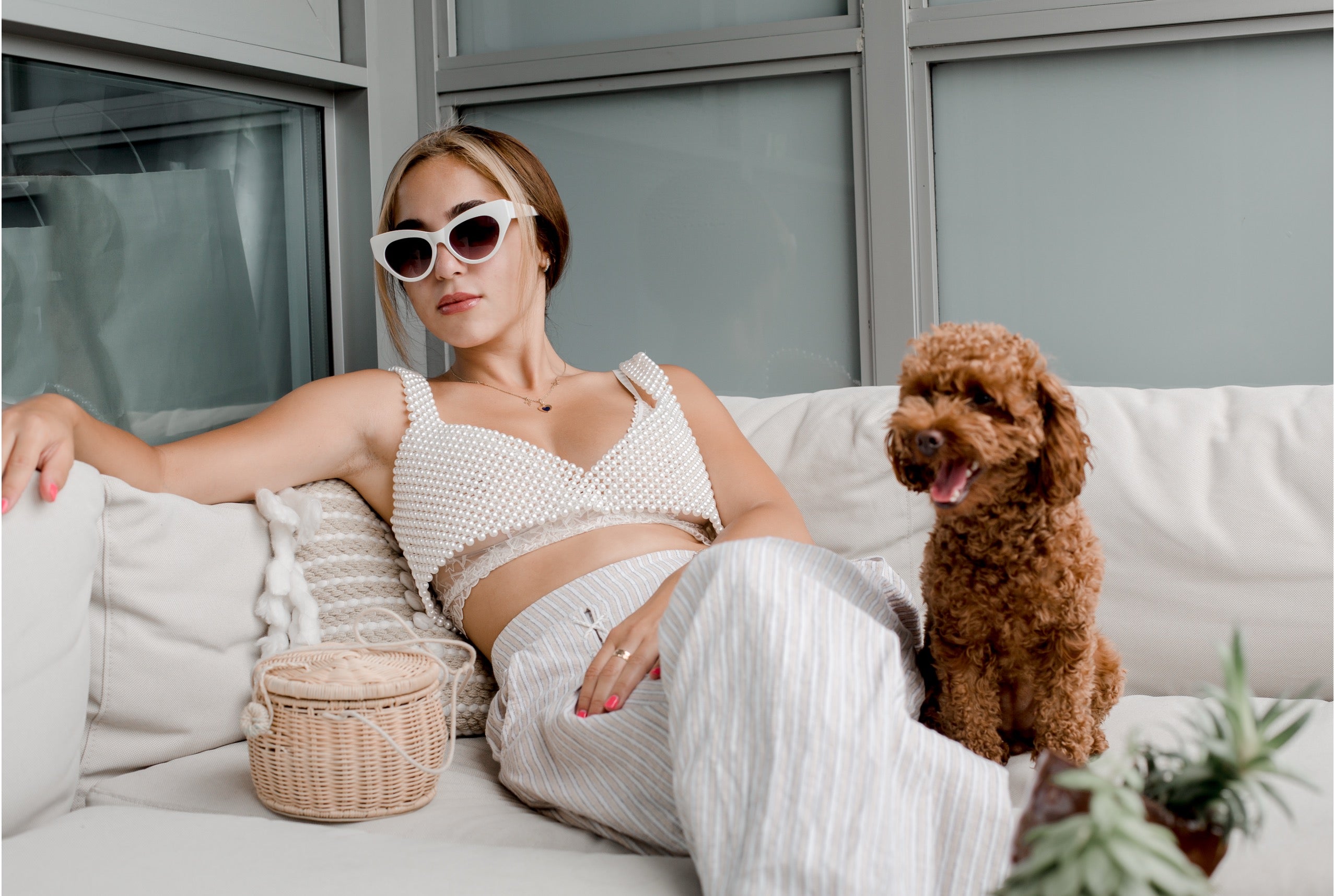A woman lounges on a white sofa wearing a matching white crochet crop top and striped white pants, with a small brown poodle beside her. She is wearing white sunglasses, and a woven basket bag sits on the couch next to her.