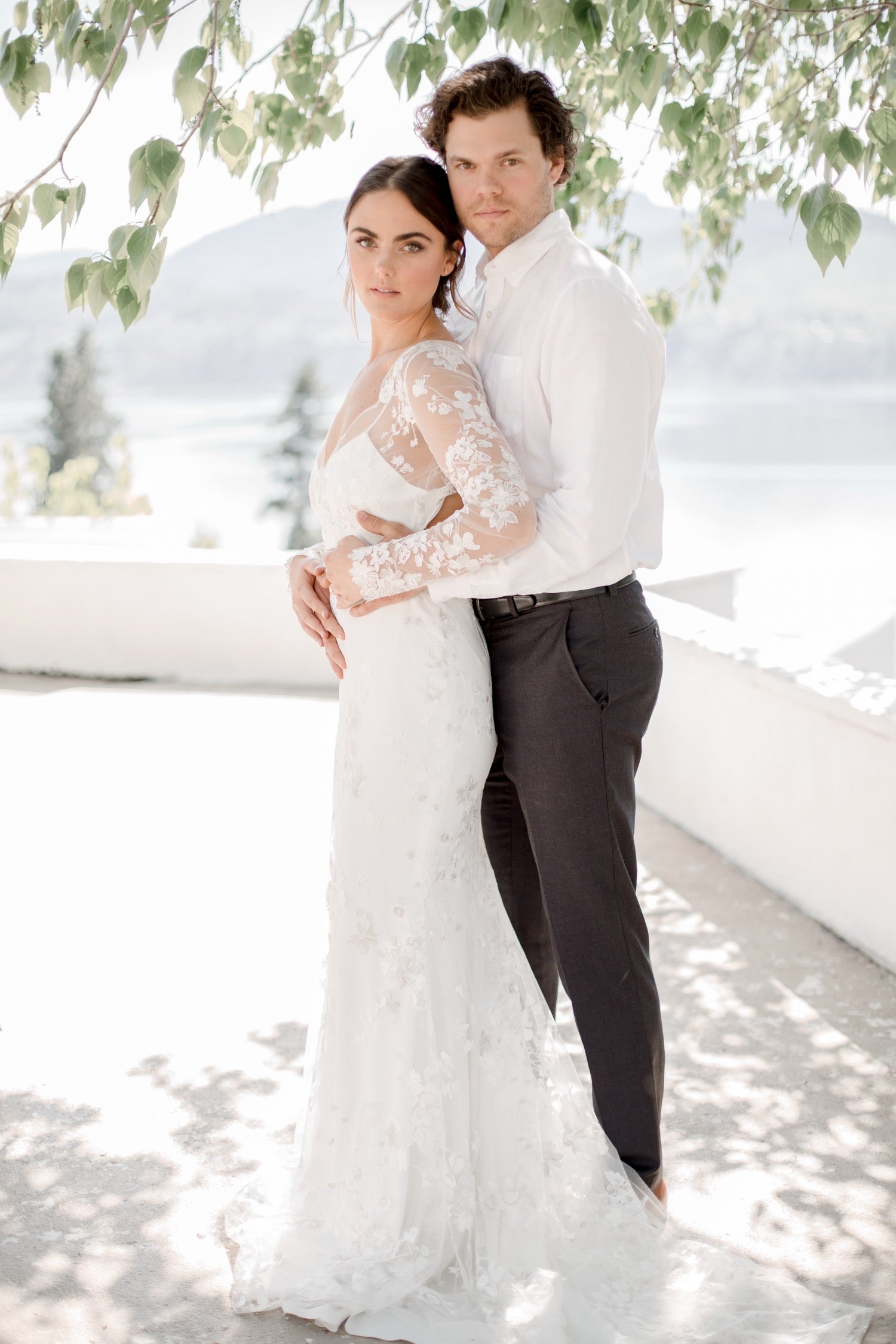 Bride and groom embracing under leafy branches with snow-capped mountains behind them
