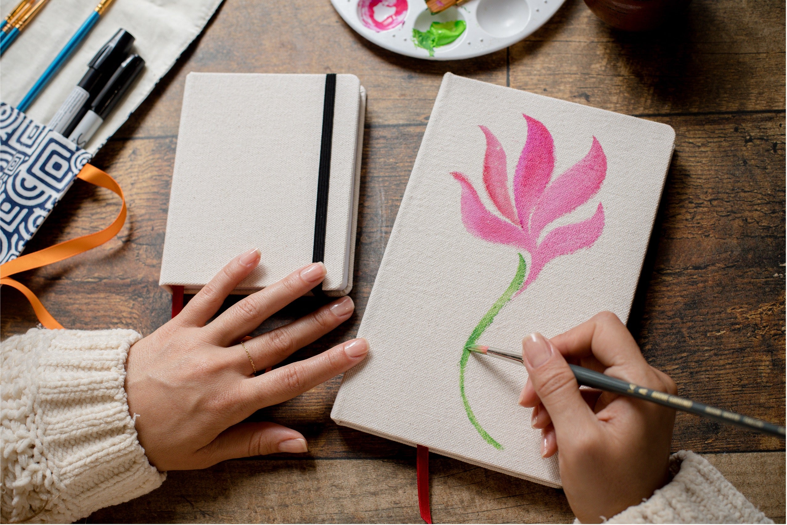 A top-down view of a person's hands painting a pink tulip on a beige linen notebook with a paintbrush, next to another closed notebook on a wooden table with art supplies.