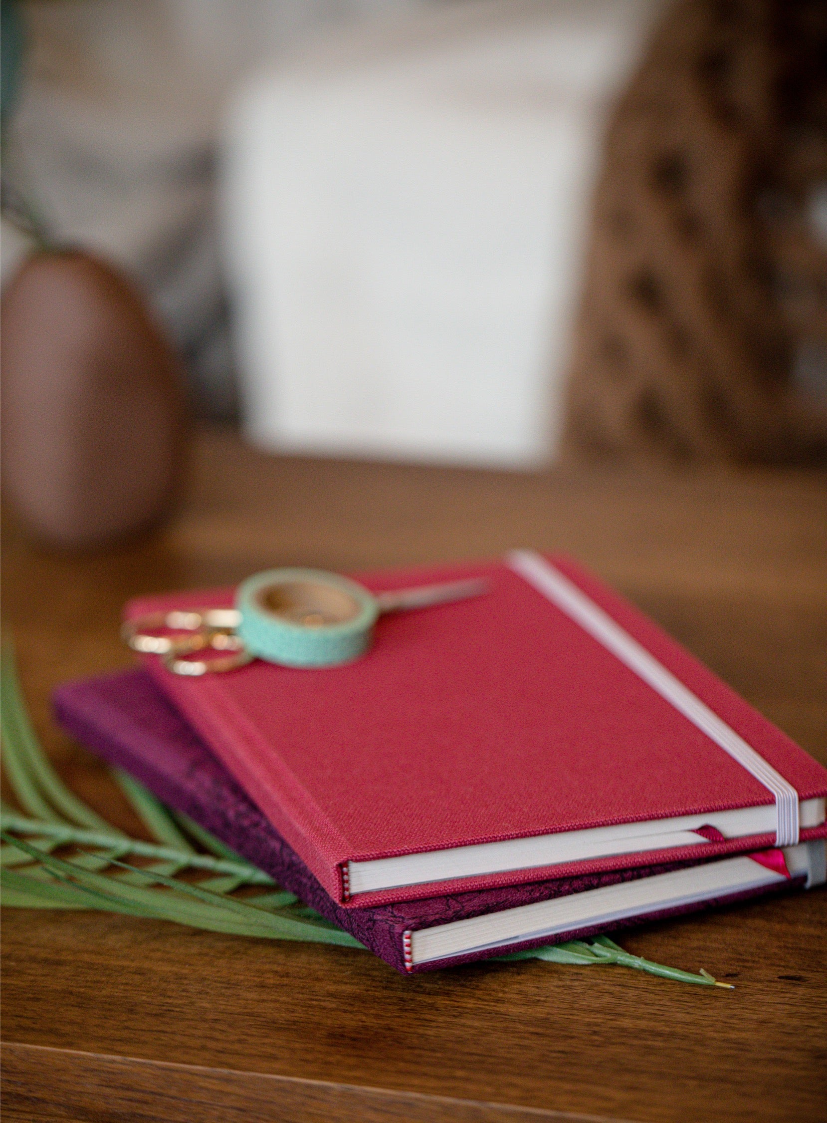 Two hardcover notebooks stacked on a wooden table, with a pink top notebook and a purple bottom notebook, decorated with a mint green washi tape roll and gold scissors.