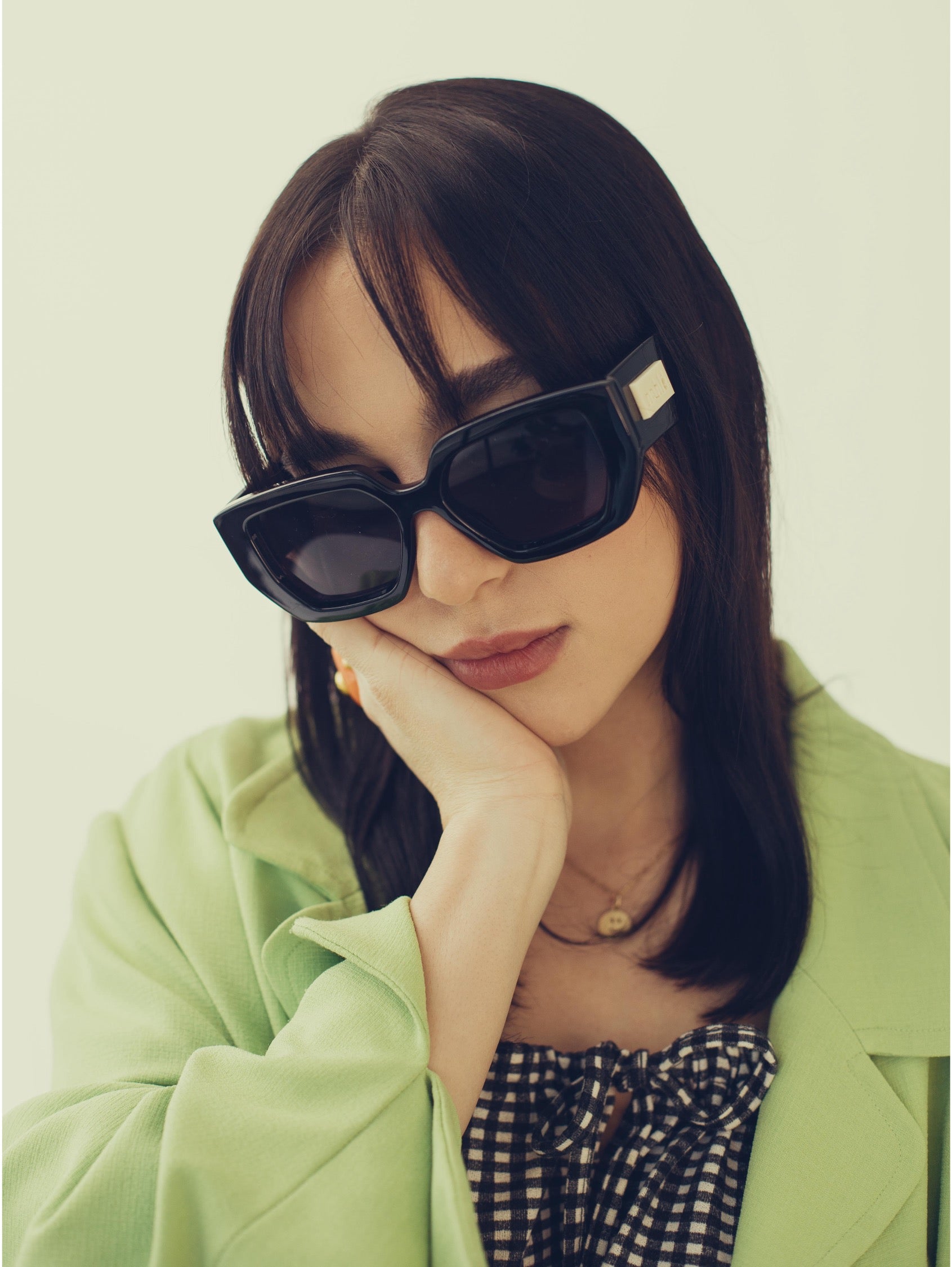 A woman wearing black oversized square sunglasses with a silver rectangular accent on the temple, resting her head on her hand. She is wearing a light green blazer and a black-and-white gingham top against a solid pale green background.
