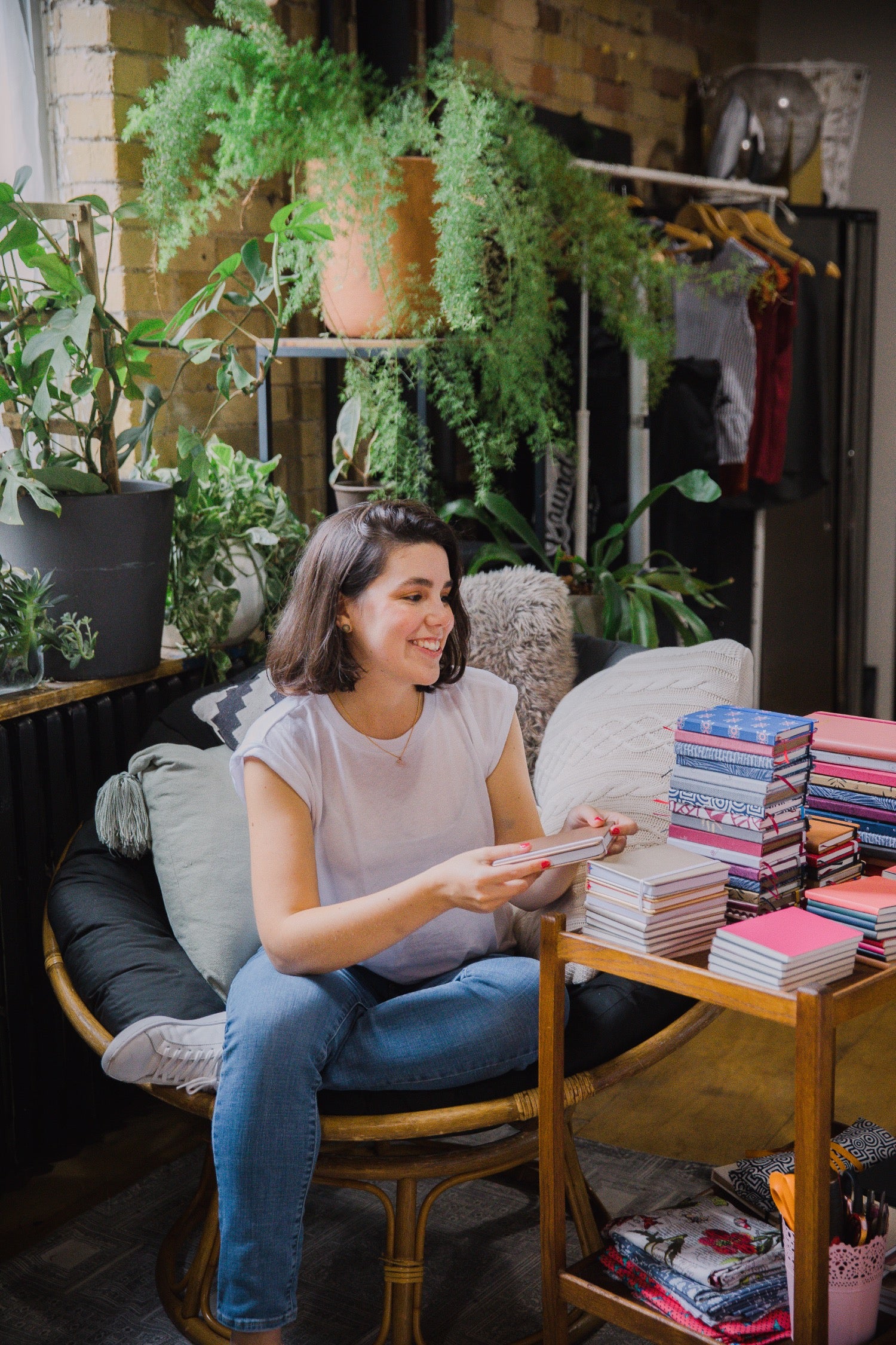 A woman sits in a round rattan lounge chair in a cozy, plant-filled room, holding a stack of colorful books or magazines. A wooden side table next to her is stacked with more books.