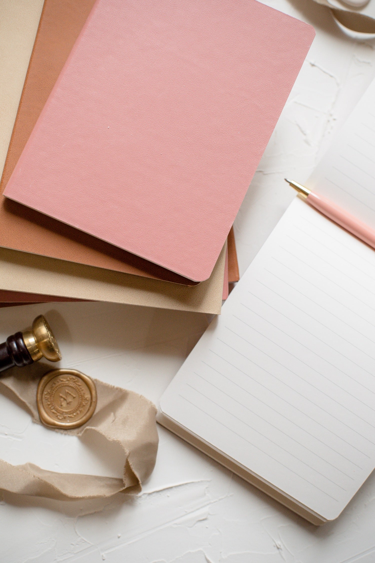 A styled flat-lay of stationery on a white textured surface, featuring a closed dusty pink hardcover notebook, an open lined notebook with a pink pen, a gold wax seal, and a gold seal stamp.