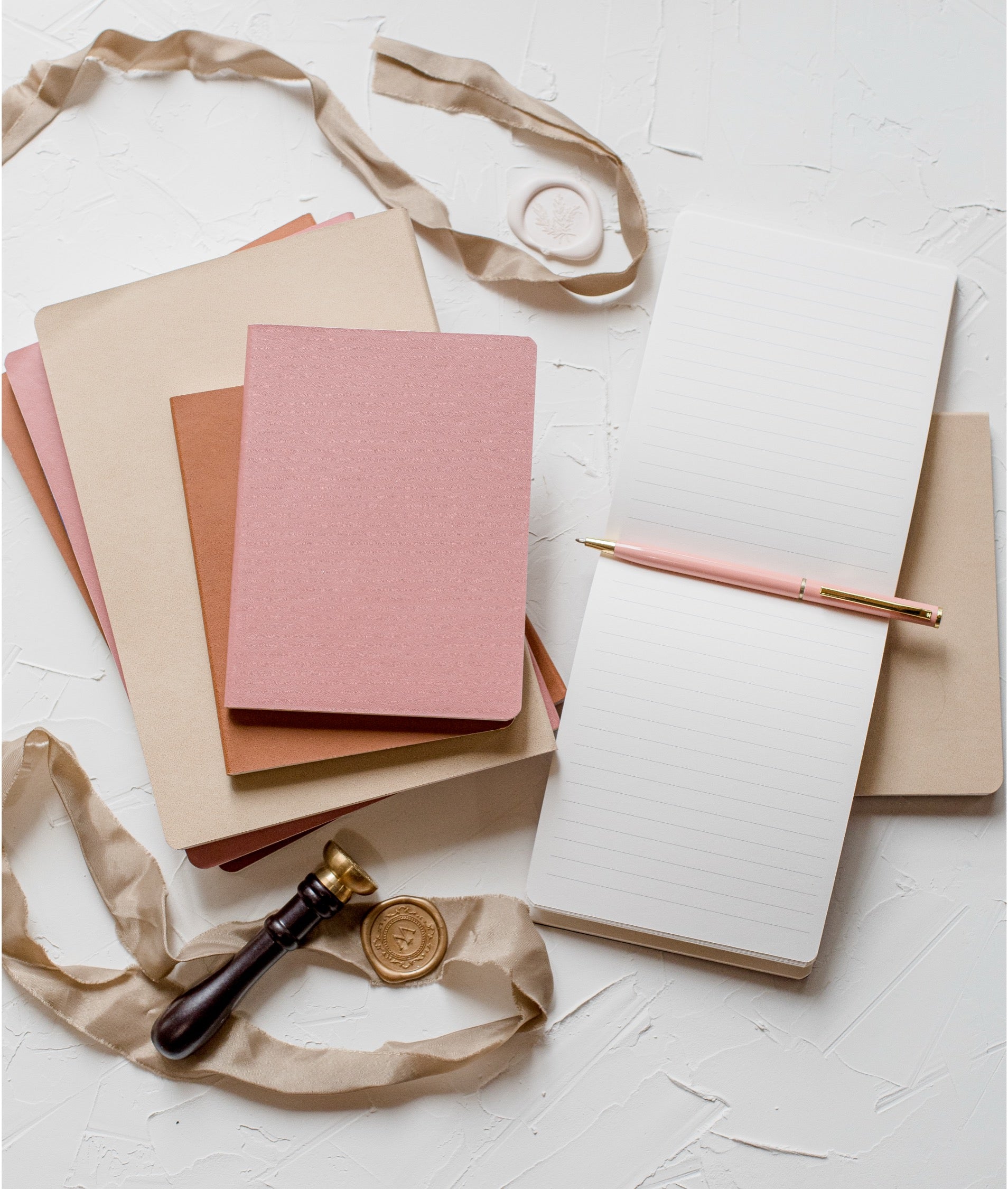 A styled flat-lay of stationery items on a white textured background, including a stack of notebooks in beige, cream, and dusty pink, an open lined notebook with a pink pen, a pink wax seal, a wax stamp with a gold seal, and beige ribbon.
