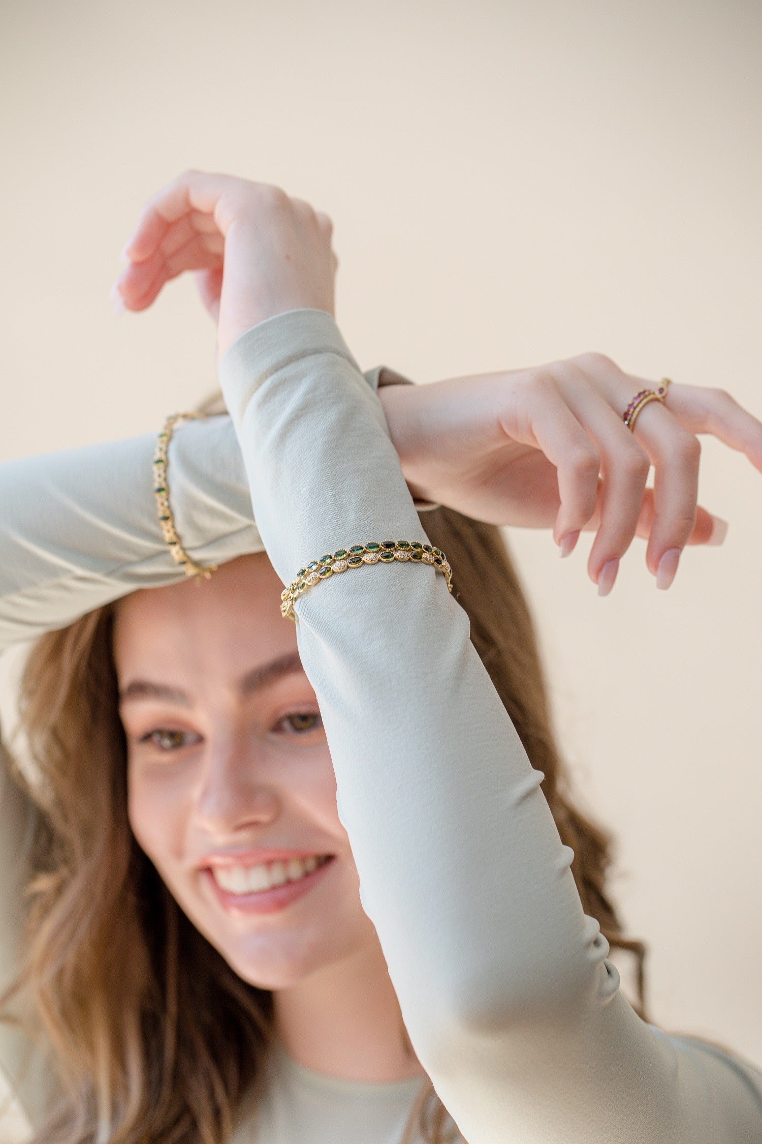 A woman wearing a gold chain bracelet and matching gold ring, posing with her arms crossed against a soft beige background.