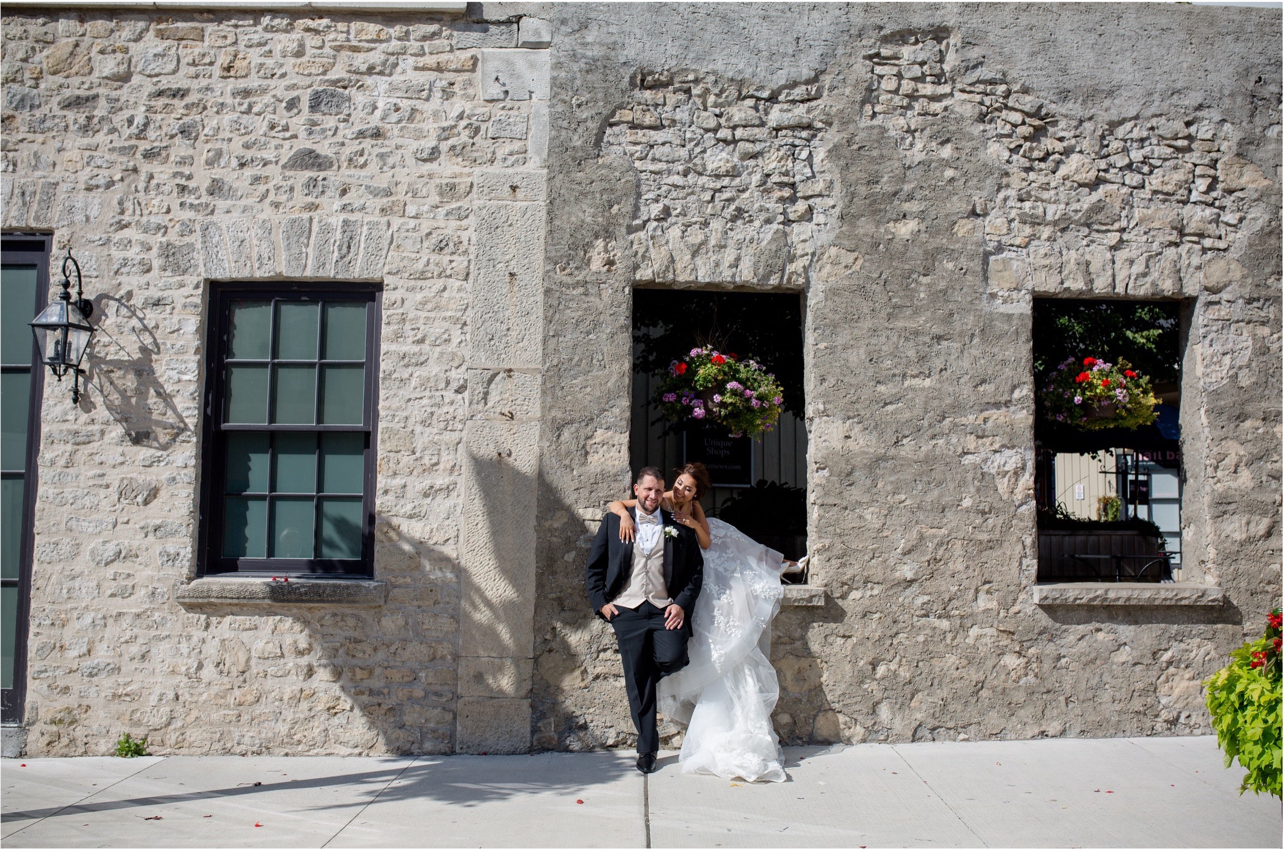 A bride in a white wedding gown and a groom in a black tuxedo stand together in front of a rustic stone building with dark-framed windows and hanging flower baskets.