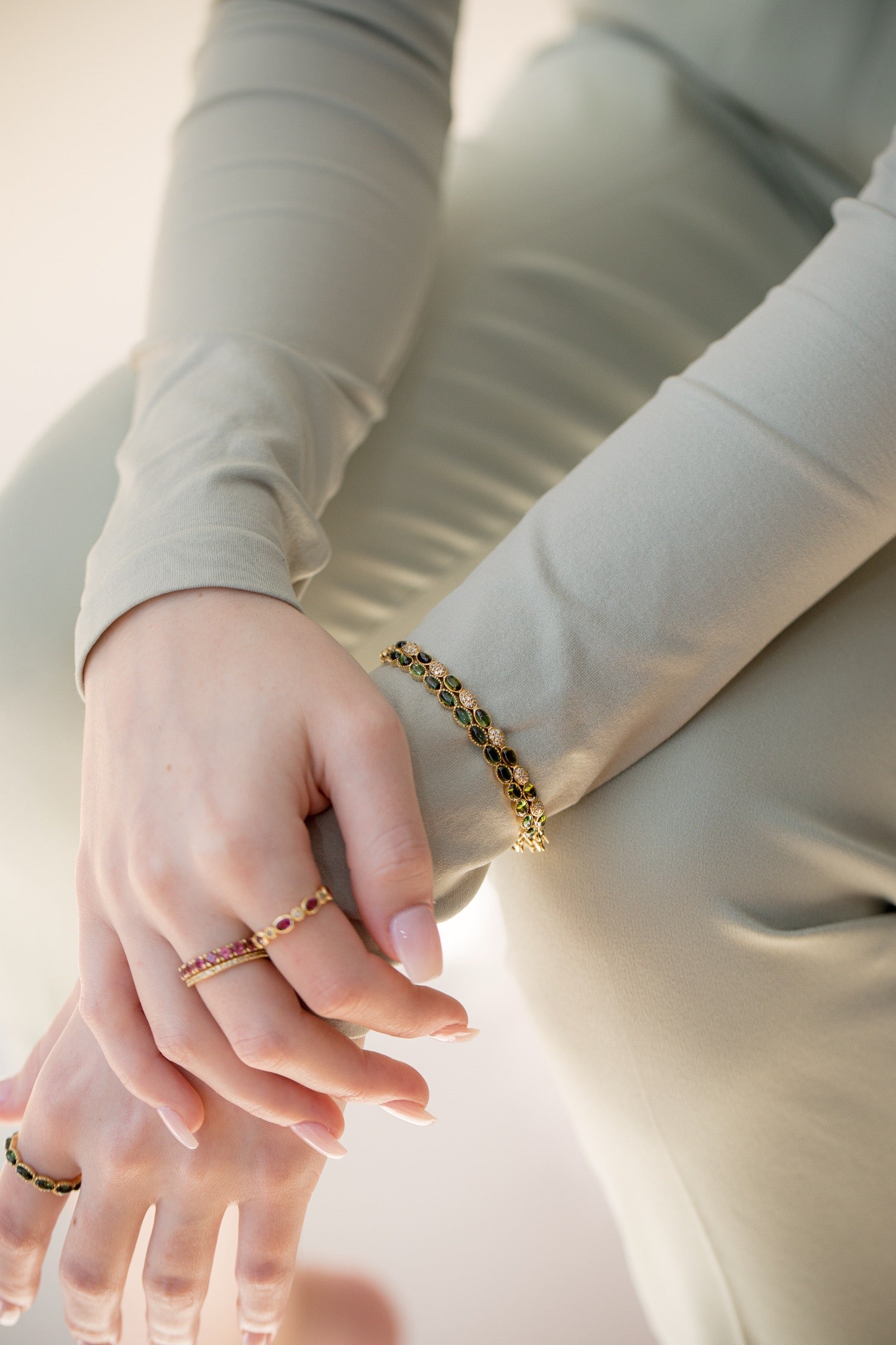 A close-up lifestyle shot of a person wearing a delicate gold bracelet with small dark green and red stones on their wrist, along with matching rings on their fingers, set against a soft beige background.