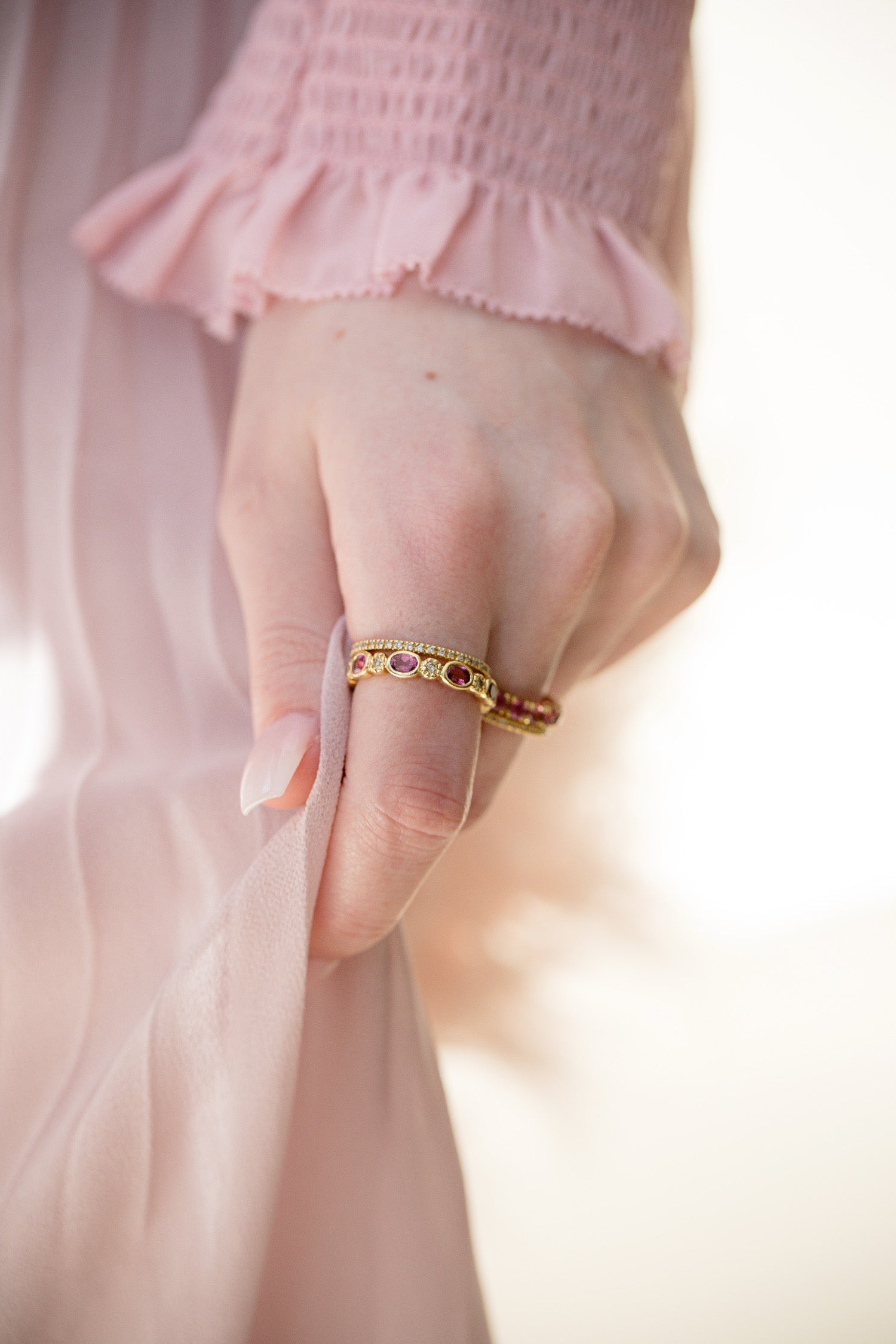 A close-up of a hand wearing two delicate gold rings, one adorned with small red gemstones and the other with clear stones, while holding a soft pink fabric.