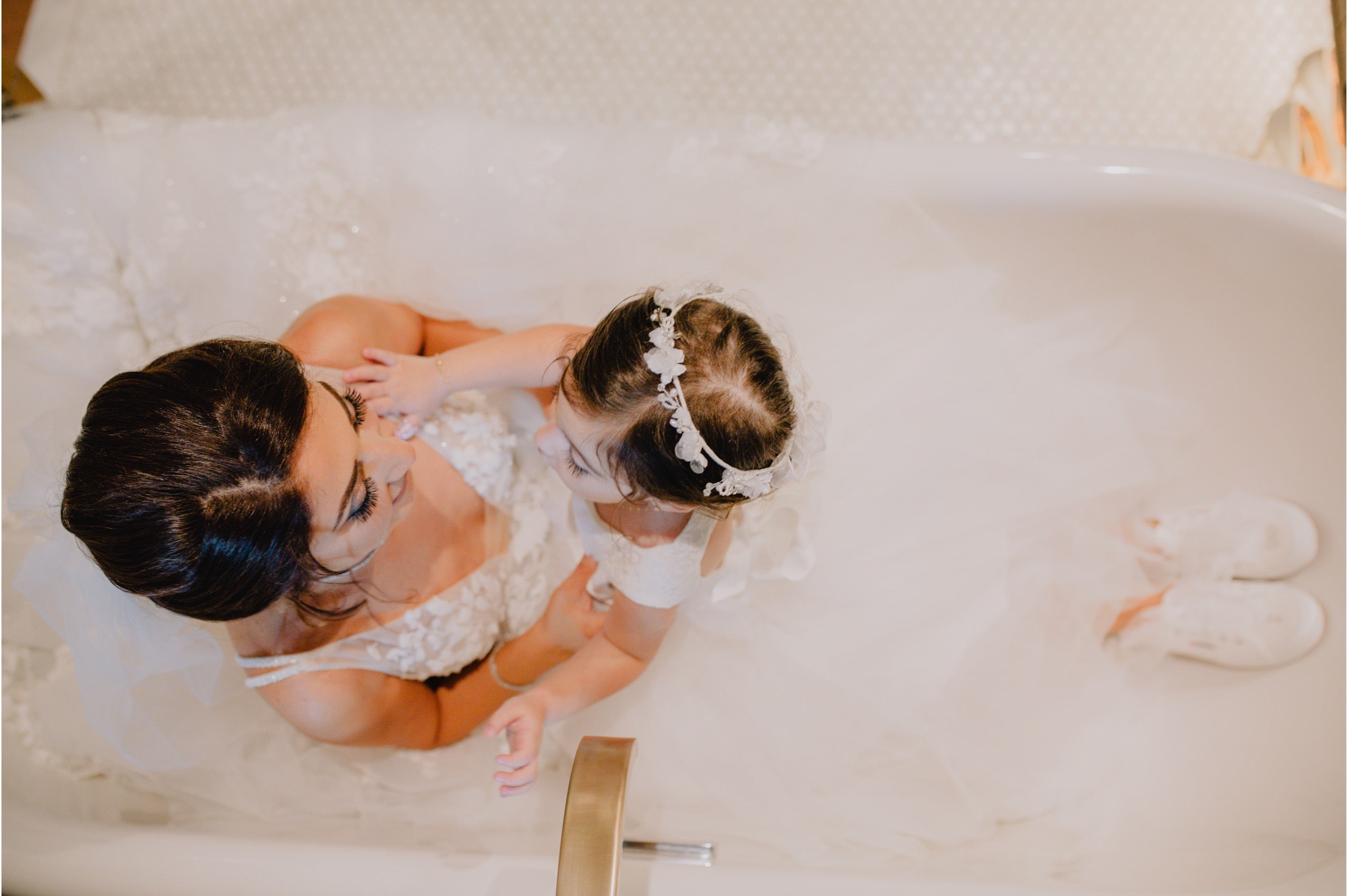 A woman and a young child wearing a white lace bridal veil and floral headpiece sit together in a bubble bath, viewed from above. The woman wears a white lace dress, and white slipper-like shoes are visible on the tub edge.