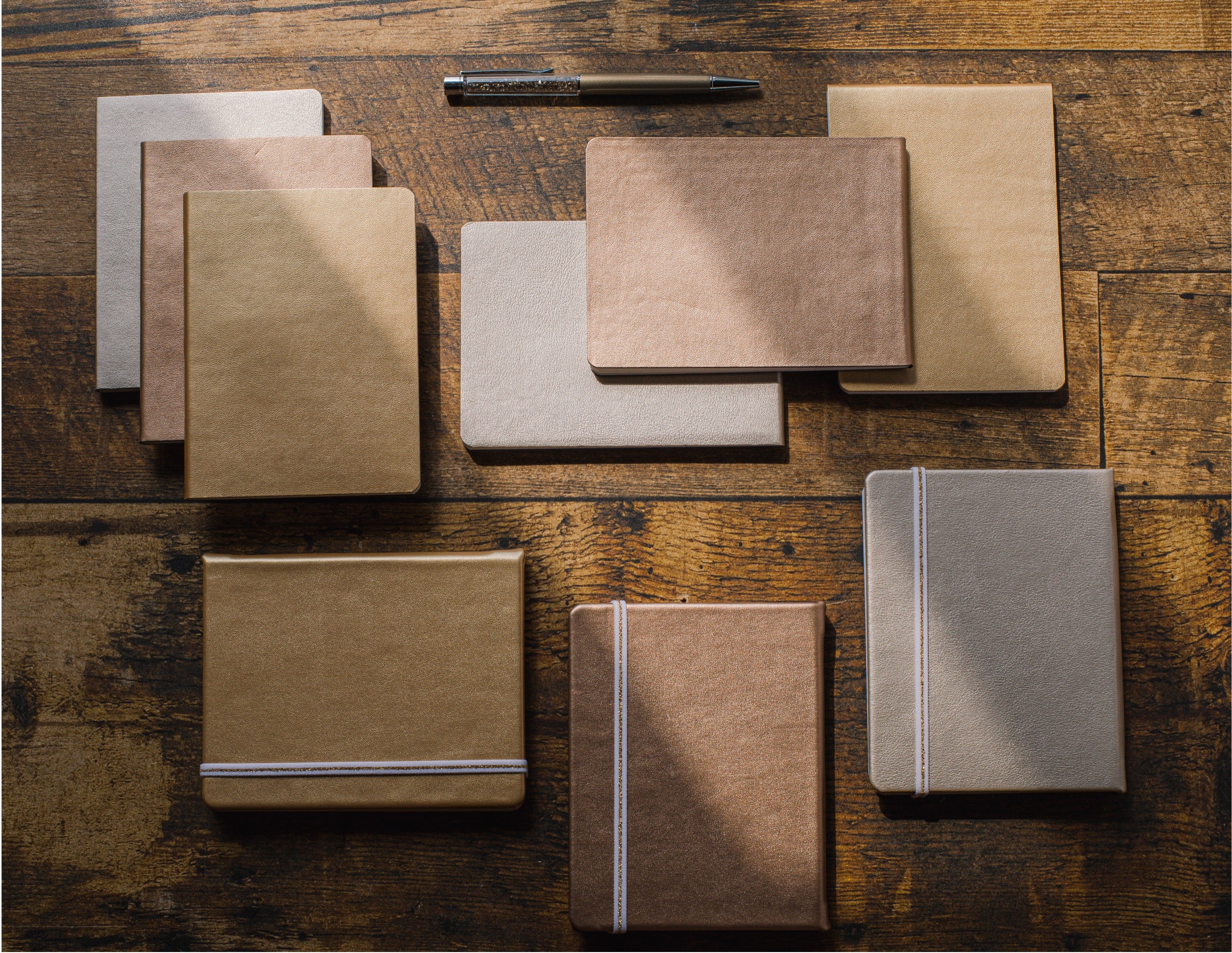 A collection of minimalist notebooks and journals in neutral earth tones, including kraft paper, beige, brown, and gray, arranged on a rustic wooden table with a pen above them.