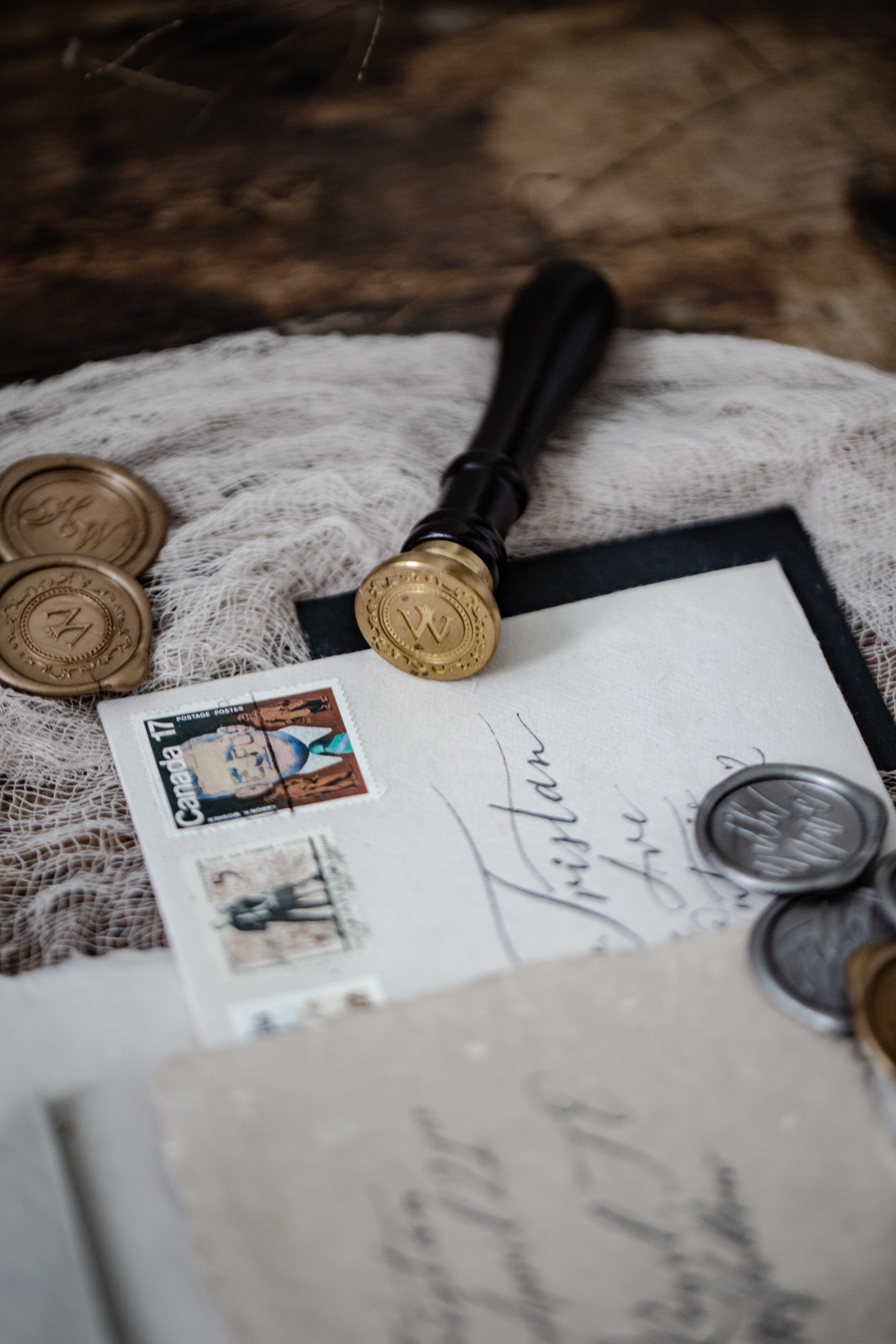 A vintage-style wax seal stamp with a black handle and gold seal face rests on a white card with handwritten script and postage stamps, surrounded by old coins and a rustic wooden surface.