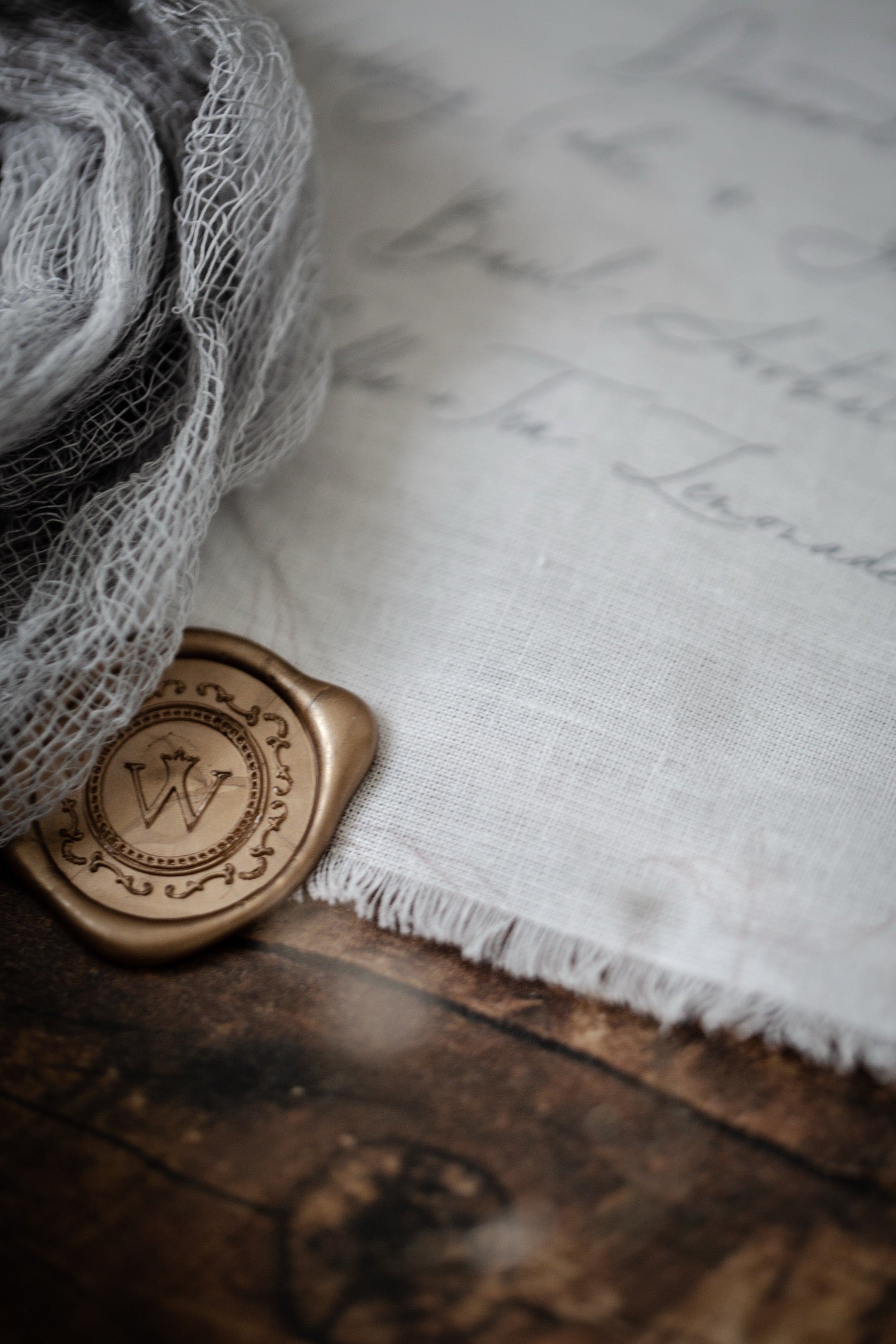A close-up styled product shot showing a rolled piece of gray gauze fabric next to a bronze wax seal stamp with an engraved monogram, placed on a rustic wooden surface with white frayed fabric and cursive script in the background.