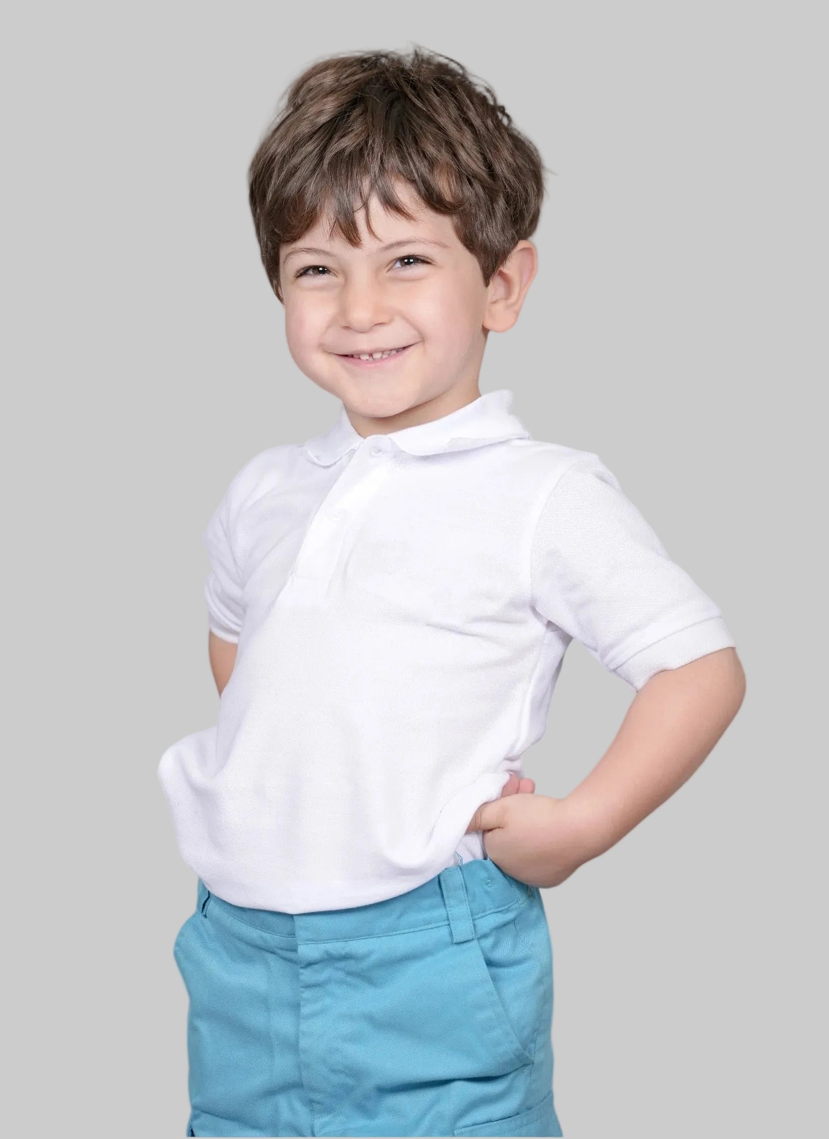 A young boy with brown hair smiling, wearing a white short-sleeve polo shirt and light blue pants, standing against a solid gray studio background.