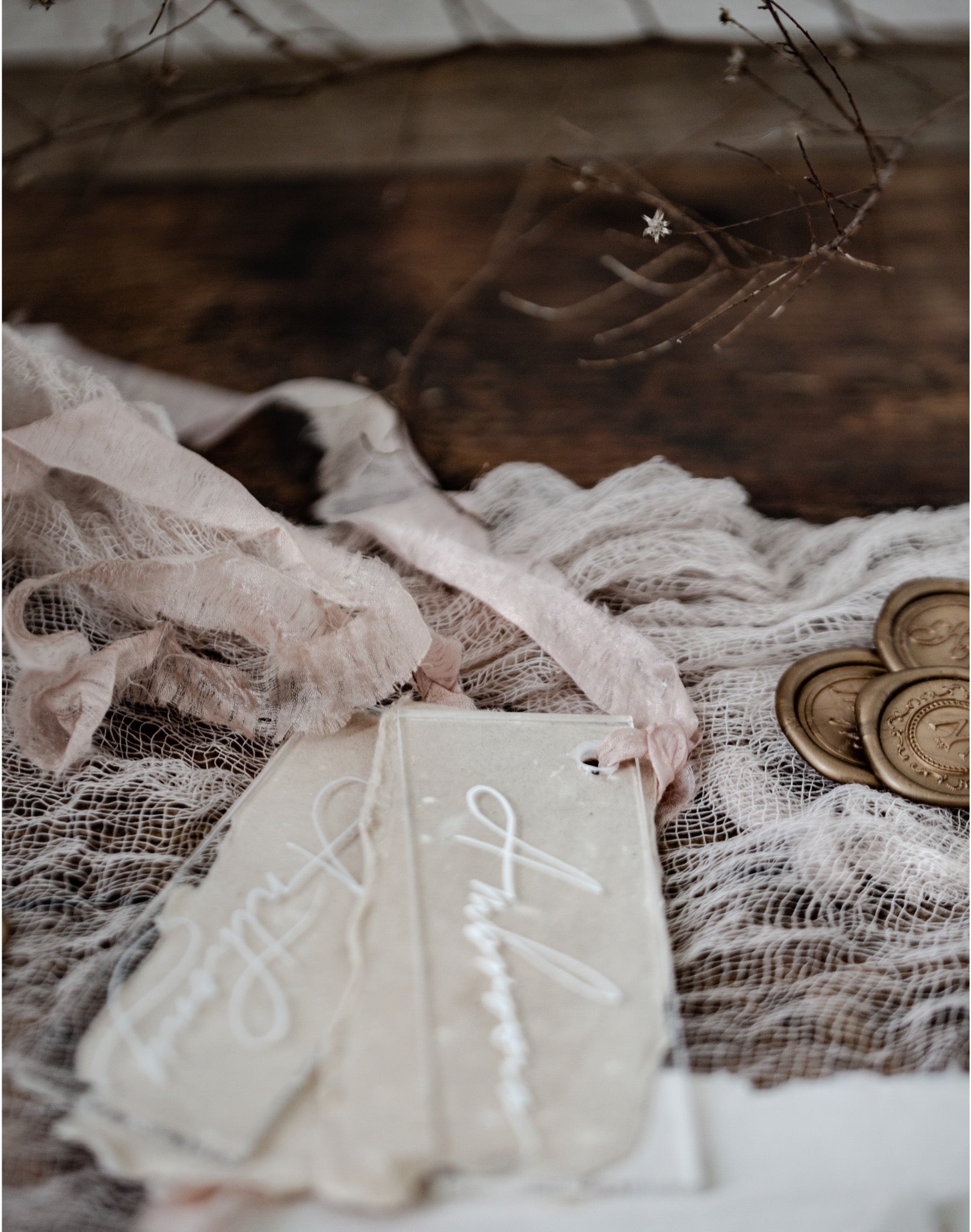 A styled flat-lay of rustic wedding favors, featuring two cream-colored paper tags with white cursive script, wrapped in a sheer blush pink ribbon, resting on a bed of white lace over a dark wooden surface, accented with gold coins and dried branches.