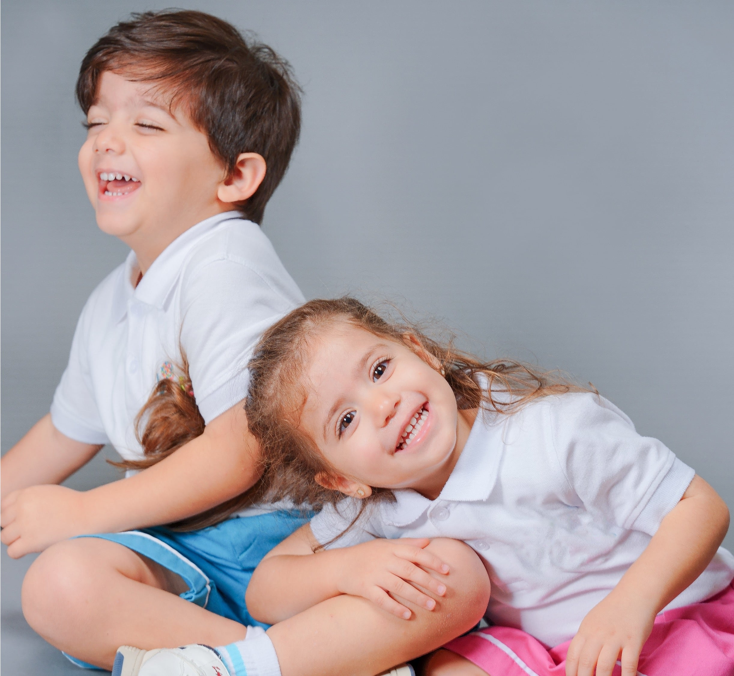 Two young children, a boy and a girl, sitting together against a solid gray studio background. Both are smiling broadly and laughing, wearing matching white short-sleeve collared shirts and colorful shorts (blue and pink).