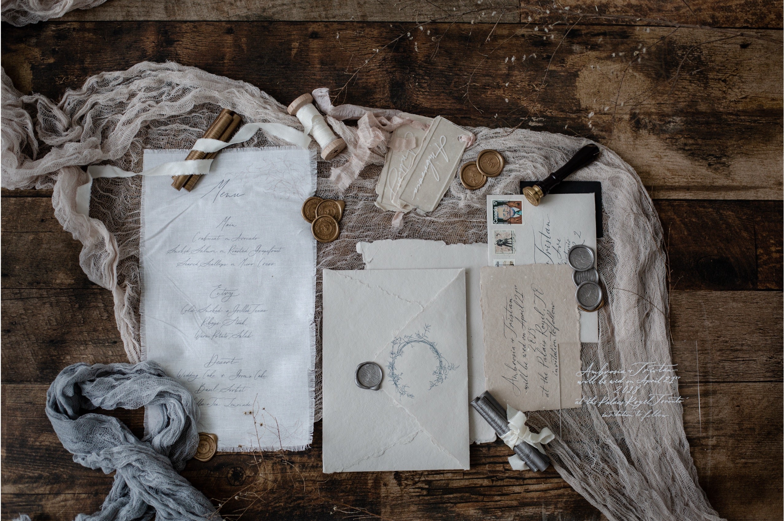 A styled flat-lay of vintage-inspired stationery on a rustic dark wood surface, including handwritten invitation cards, envelopes, old coins, a small bottle, ribbon, and a gray scarf.