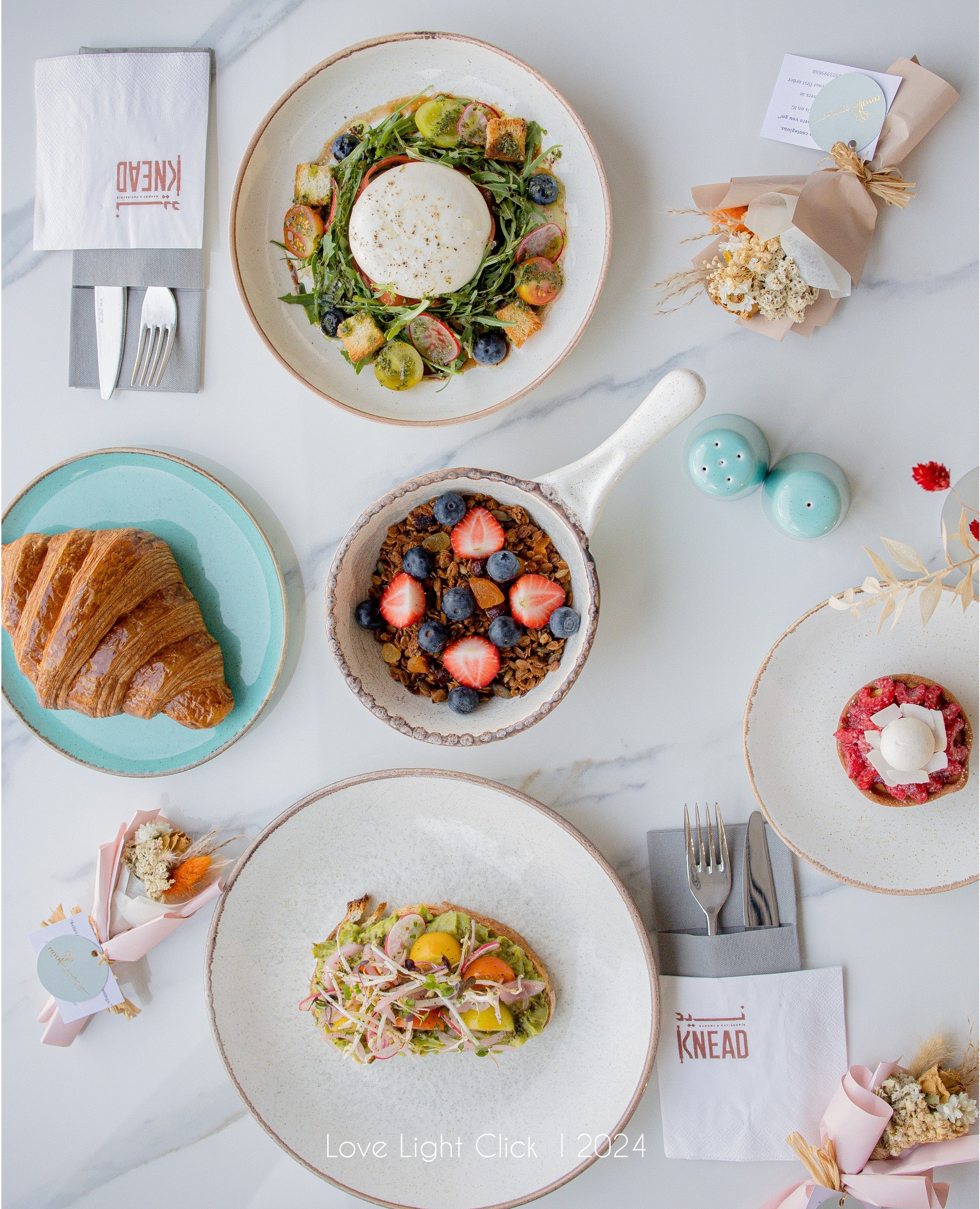 A styled flat-lay of multiple breakfast dishes on a light marble surface, including a croissant on a turquoise plate, a salad with figs and blueberries, granola with strawberries, and small pastries. Napkins with the Knead logo are visible, along with decorative flowers and pastel accents.