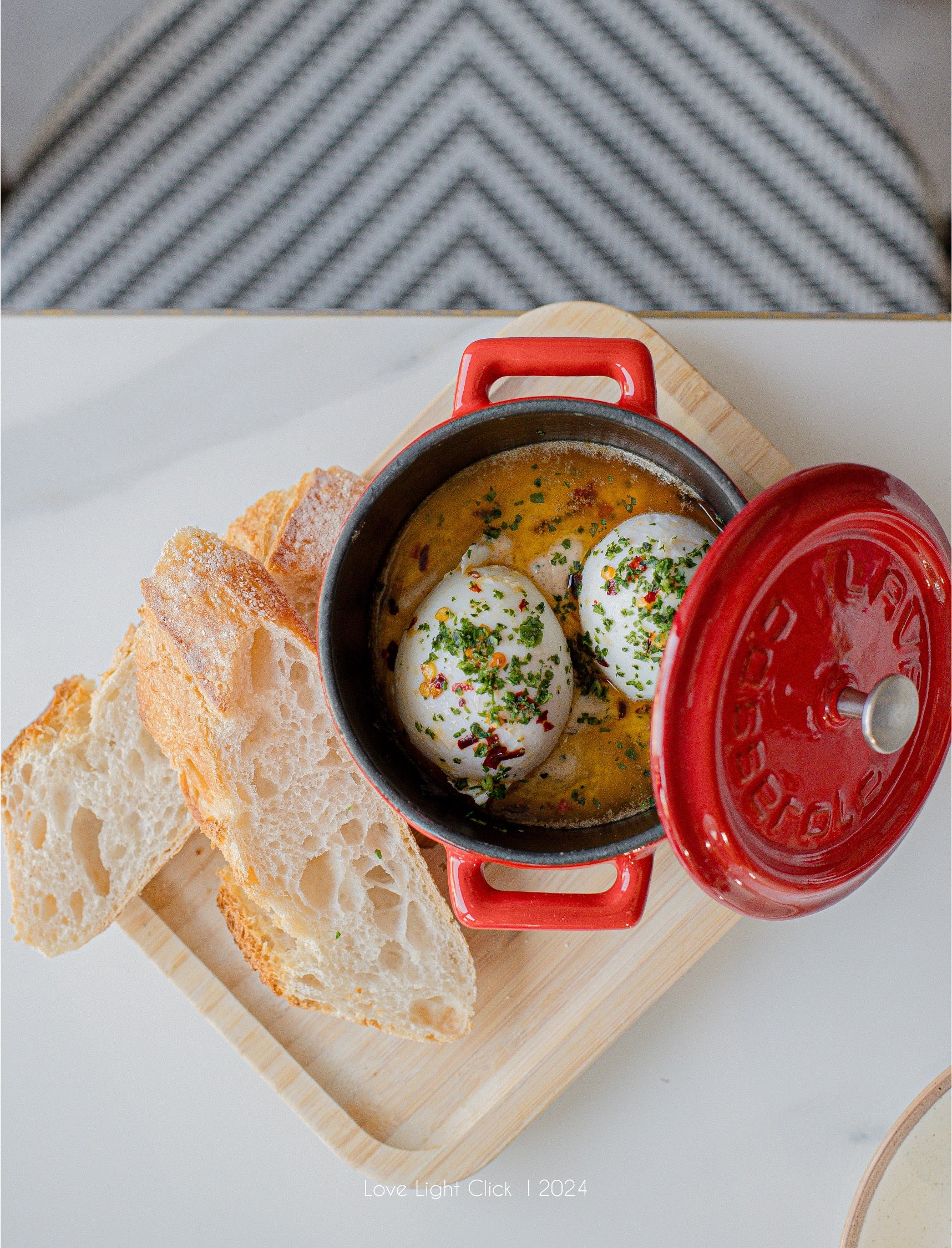 A red enameled cast iron pot filled with poached eggs in a yellow-orange sauce, garnished with chopped chives and red pepper flakes, served with slices of rustic bread on a wooden board.