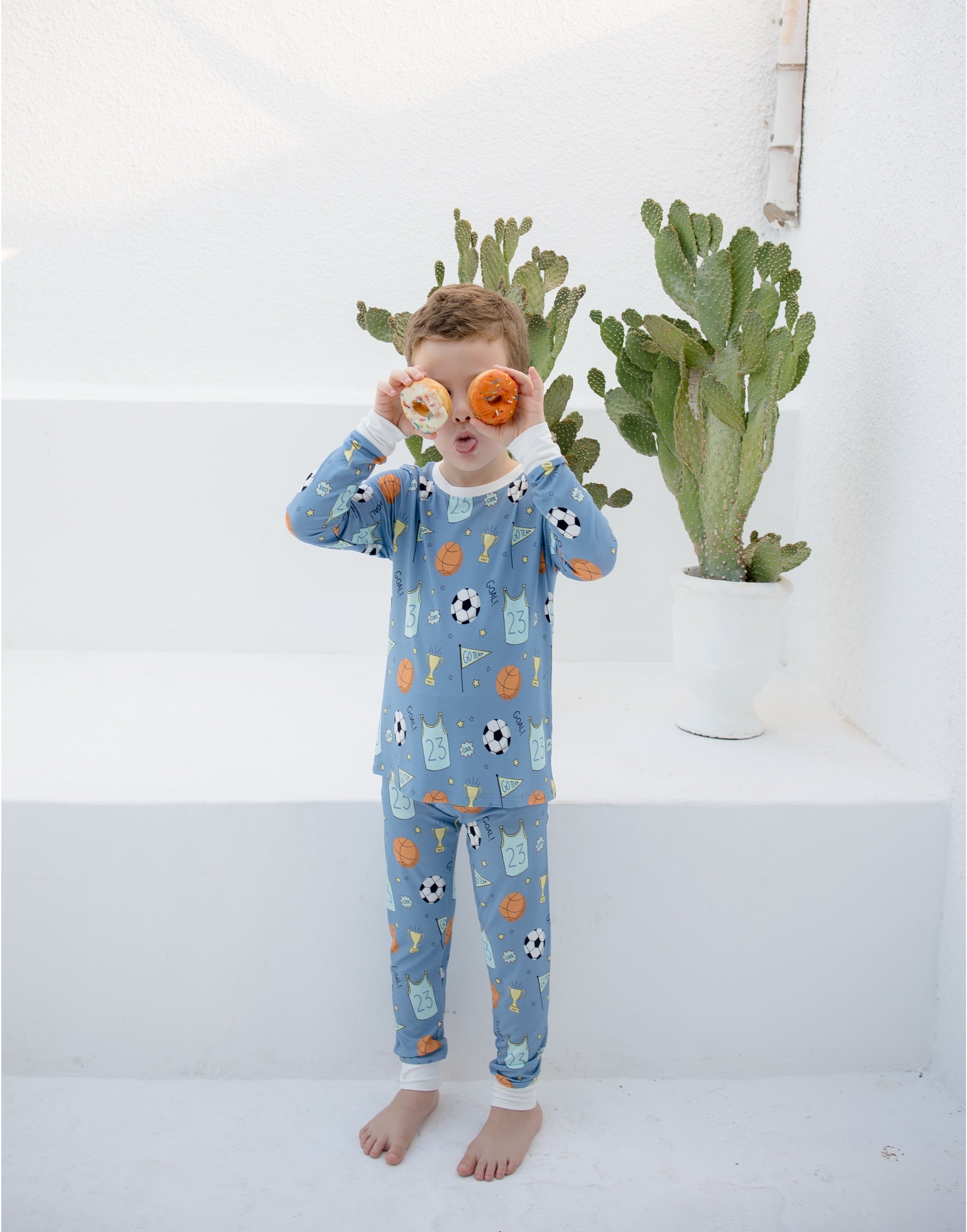 A young boy wearing a light blue pajama set with a soccer-themed print stands barefoot on white steps, holding two donuts over his eyes. Cacti are visible in the background.