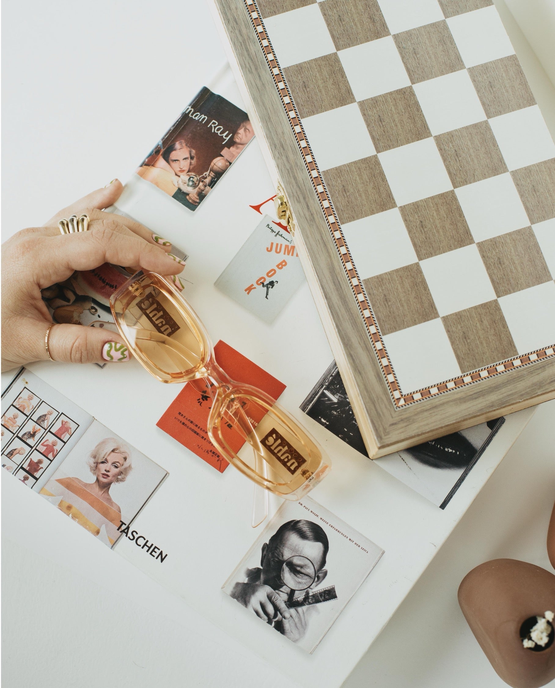 A pair of translucent amber-tinted sunglasses with a wooden 'Liberty' logo frame, held by a hand over a styled flat-lay of vintage postcards and a wooden checkerboard box.