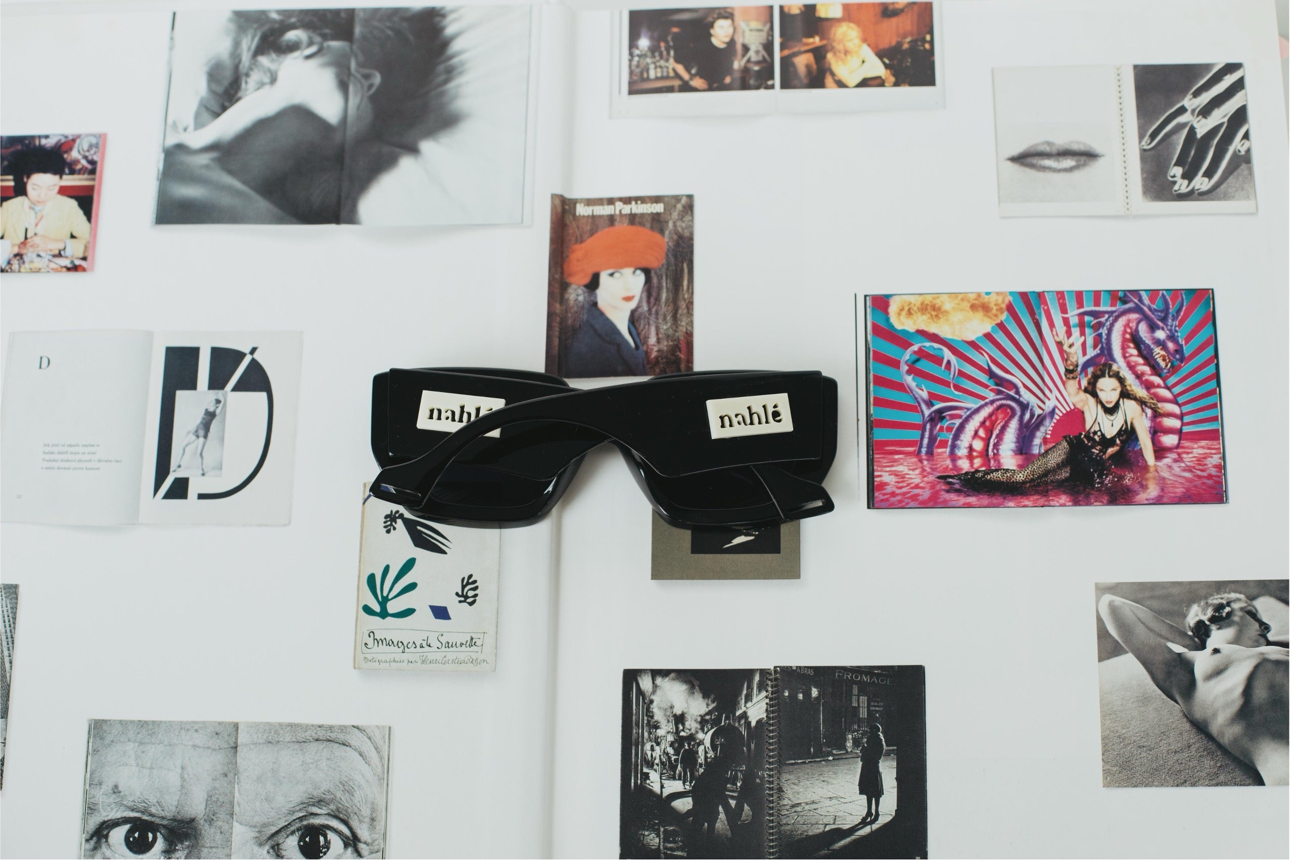 Black nablé sunglasses displayed on a white wall collage with fashion and art prints. The sunglasses feature a glossy finish with white nablé brand labels on the temples and a white hangtag.