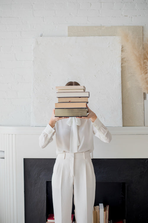 A person in a white blouse and white trousers stands in front of a fireplace holding a tall stack of books over their head, against a white brick wall and framed artwork.