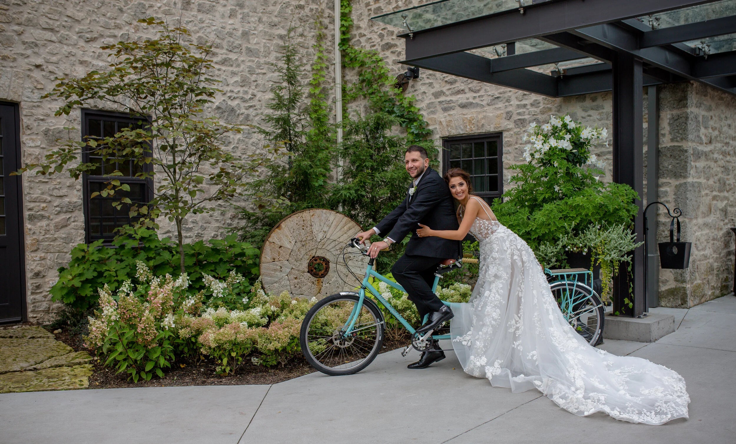 A bride in a long white lace wedding gown rides a turquoise bicycle while a groom in a black suit sits behind her, outdoors in front of a stone building with greenery and a covered entryway.