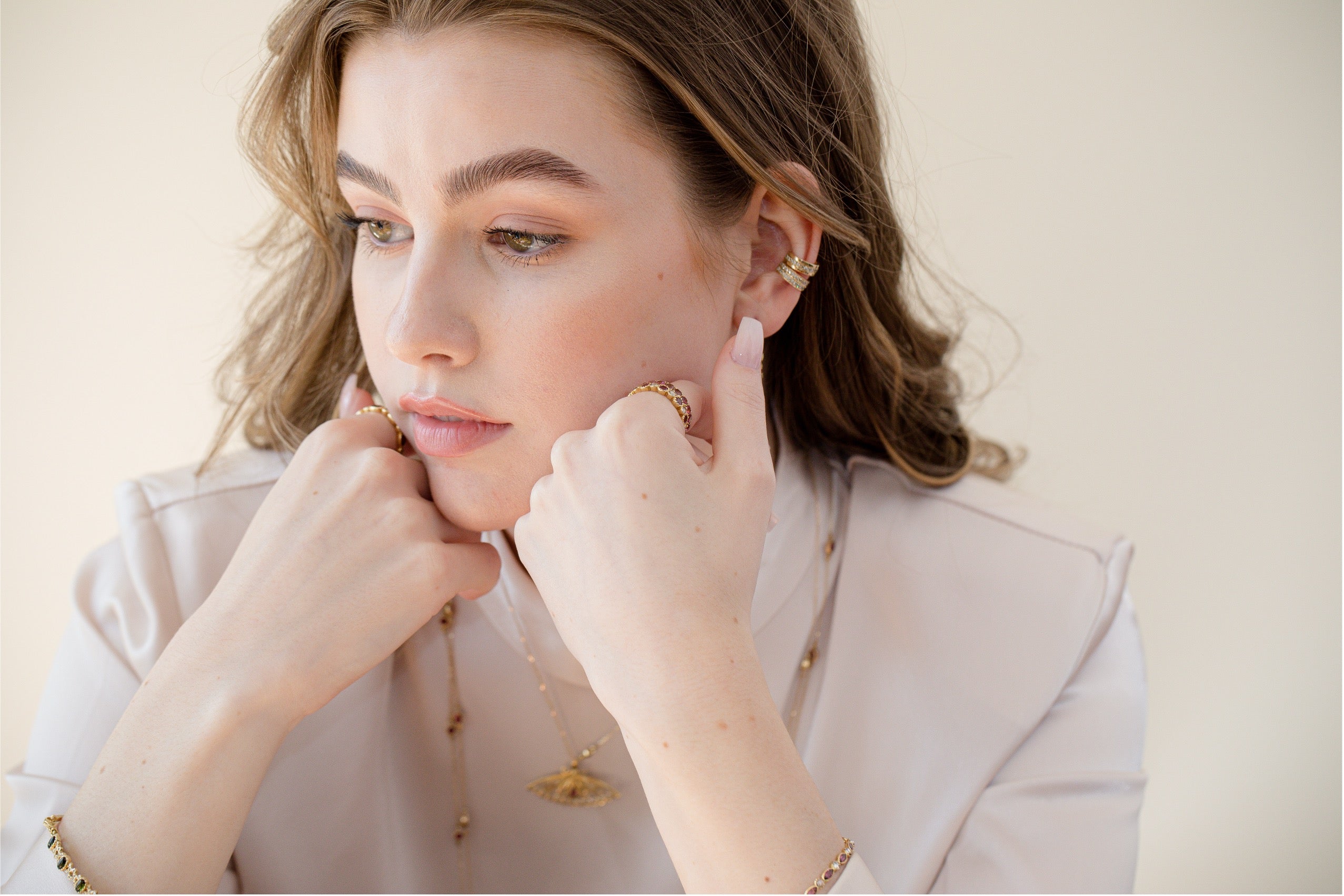 A woman with light brown hair wearing delicate gold jewelry, including a gold necklace, bracelet, and earrings, posing in a light-colored blouse against a soft beige background.