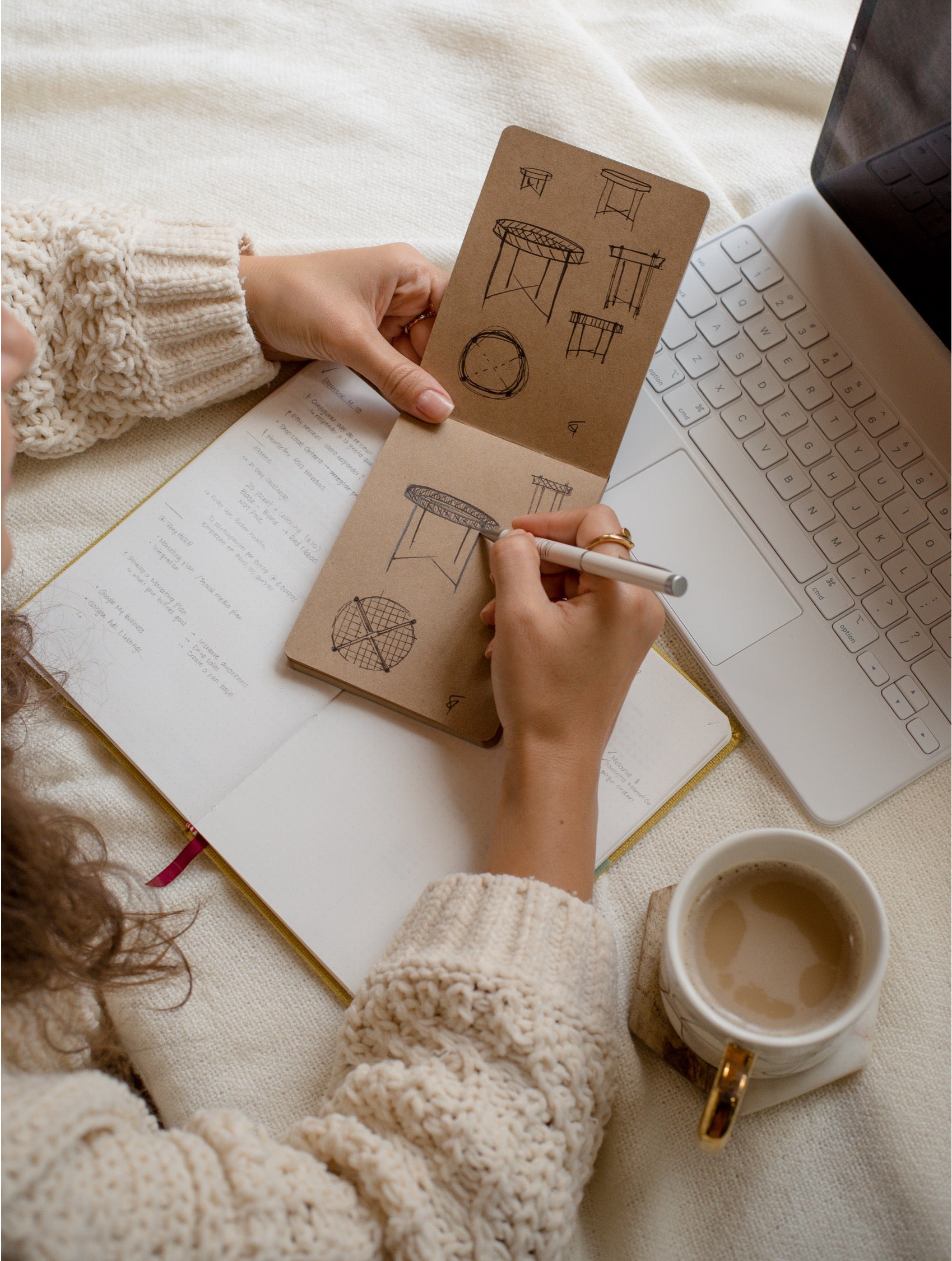 A person in a cream knit sweater sketches in a small brown notebook with a pen, styled on a cream blanket with an open laptop and a cup of coffee nearby.