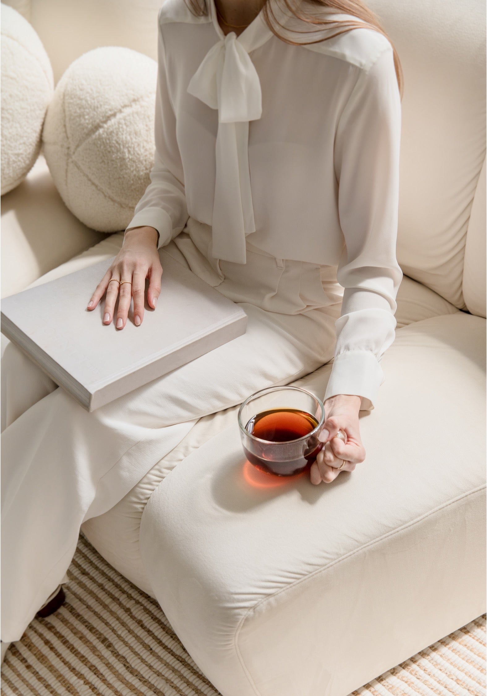 A woman wearing a cream-colored long-sleeve blouse with a bow collar and matching trousers sits on a beige sofa, holding a glass of tea and resting a book on her lap.