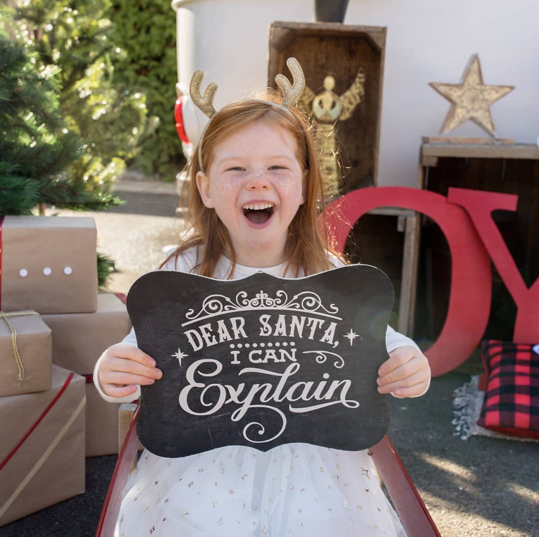 A young girl with freckles and reindeer antlers sits outdoors holding a black decorative sign that reads 'DEAR SANTA, I CAN Explain' in white script. She is wearing a white dress, with wrapped gifts, a Christmas tree, and holiday decorations in the background.