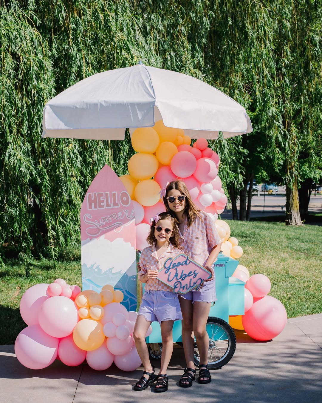Two girls posing in front of a colorful birthday party setup with a white umbrella, pastel balloon garlands, a 'Hello Summer' sign, and a 'Good Day, Girls' sign on a lawn.