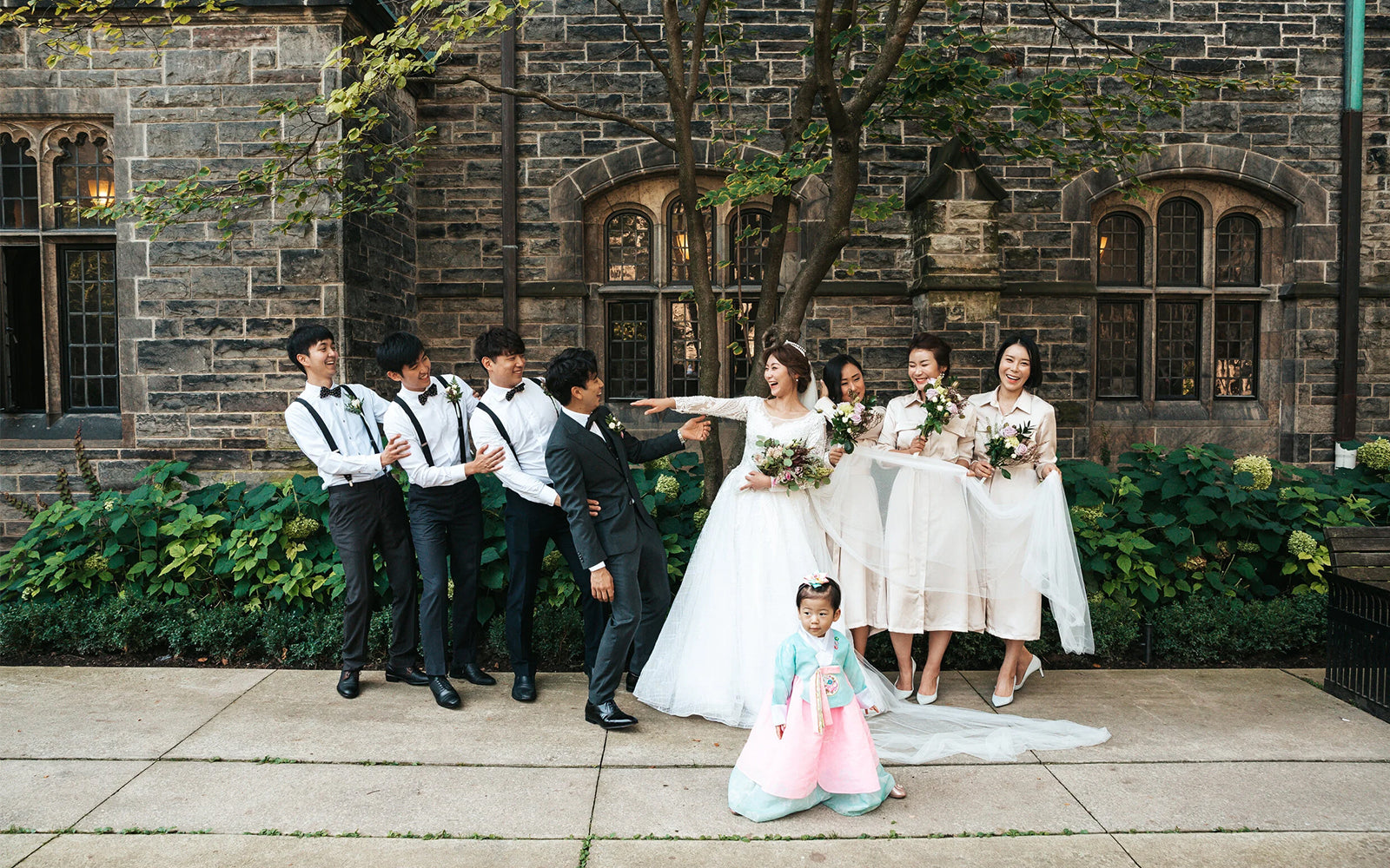 A wedding party poses outdoors in front of a stone building. The bride in a white gown holds a bouquet and points toward the groom, who is in a dark suit.