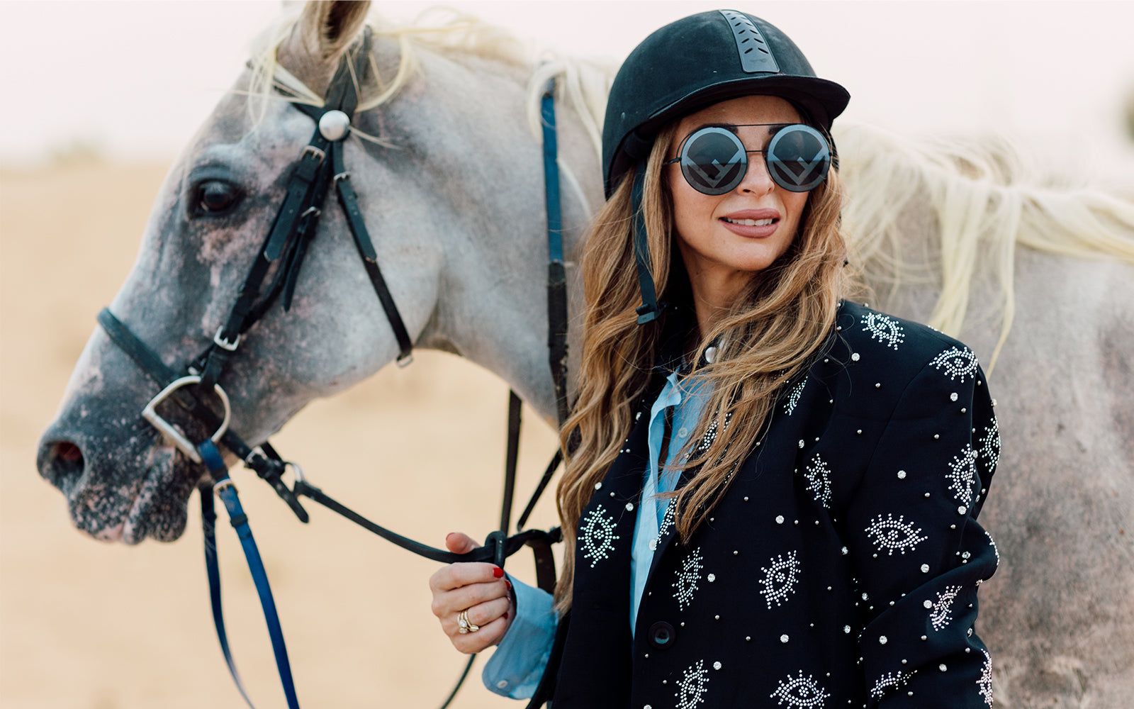A woman in equestrian attire, including a black riding helmet, round sunglasses, and a black embroidered jacket, stands next to a gray horse in a sandy outdoor setting.