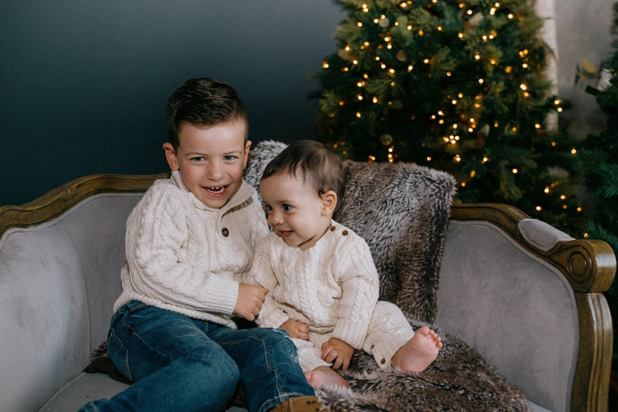 Two children sitting on a gray upholstered wooden sofa in a holiday setting, with a lit Christmas tree in the background. The child on the left wears a cream cable-knit sweater and blue jeans, while the child on the right wears a matching cream cable-knit romper.