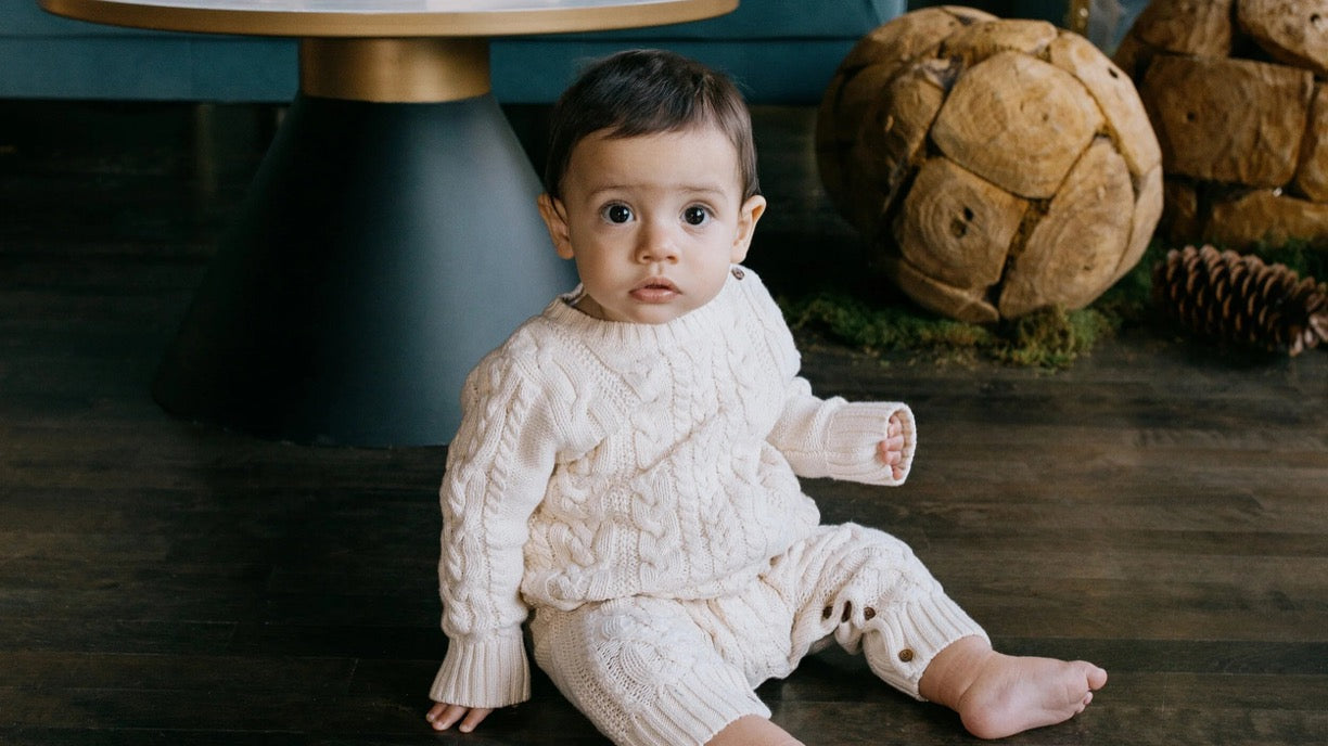 A baby wearing a cream cable-knit sweater and matching knit pants sits on a dark wooden floor in a styled indoor setting with decorative dried flowers and a table in the background.
