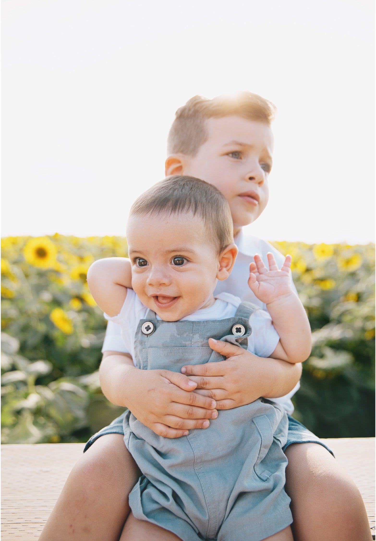 A baby in light blue denim overalls and a white t-shirt sits on a man's lap outdoors in a sunflower field. The man wears matching denim overalls and a white t-shirt.