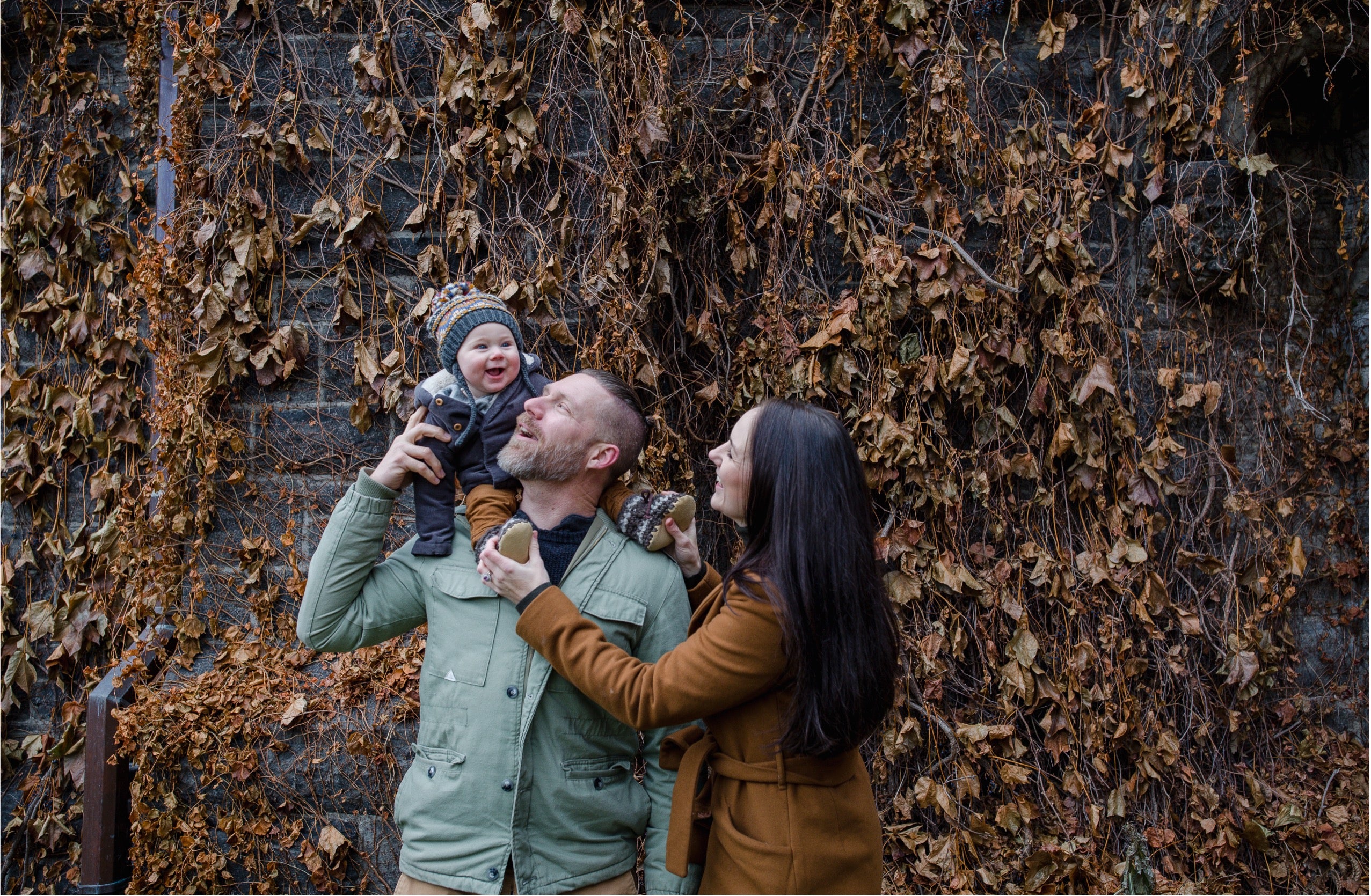 A family of three posing outdoors against a wall covered in dry brown autumn ivy. A man in a sage green jacket holds a baby wearing a knit beanie and a smiling mother in a brown coat looks on.