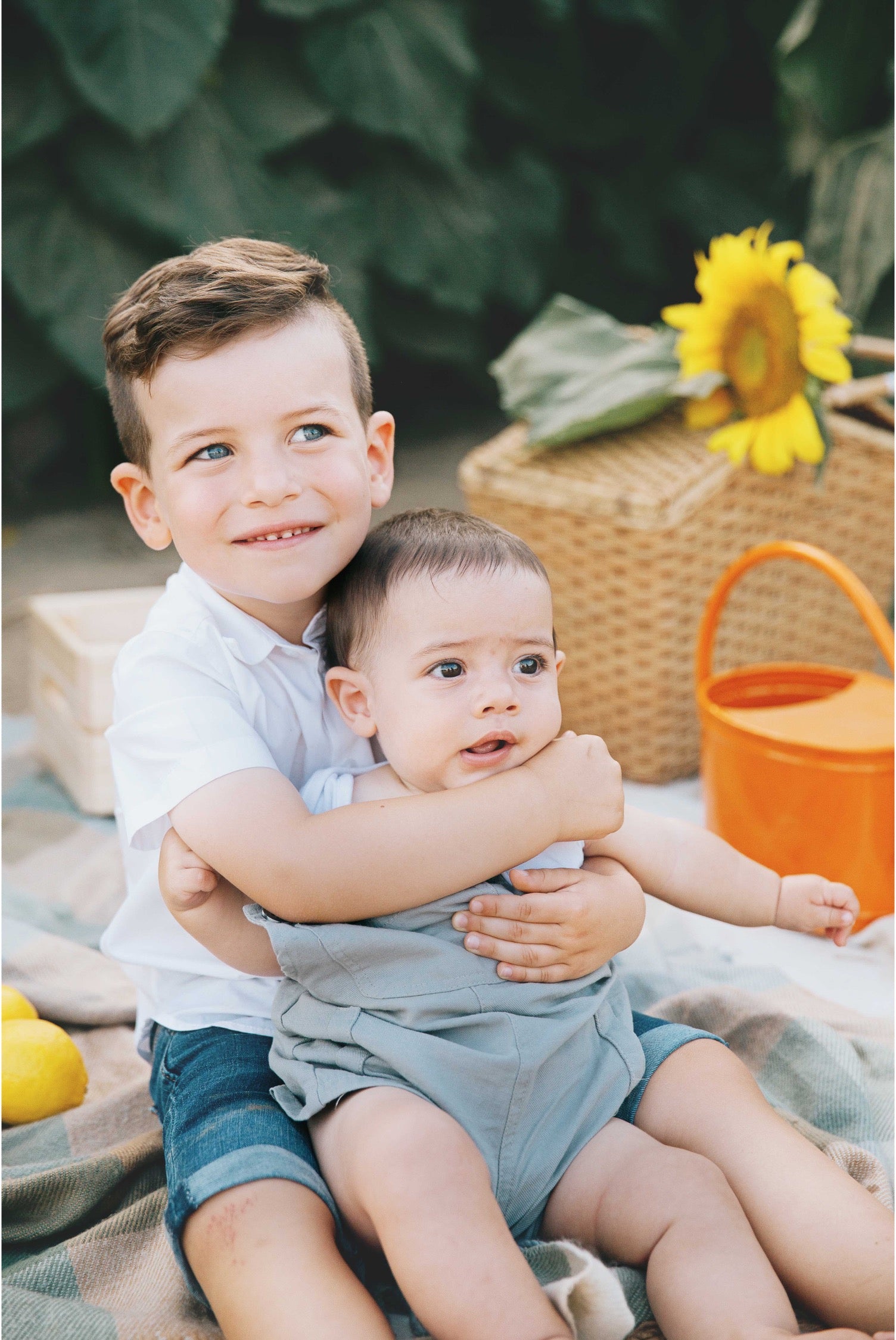 A young boy in a white polo shirt and denim shorts hugs a baby wearing a light blue-gray romper and matching shorts, sitting on a plaid blanket outdoors with a wicker basket, sunflower, and orange watering can in the background.