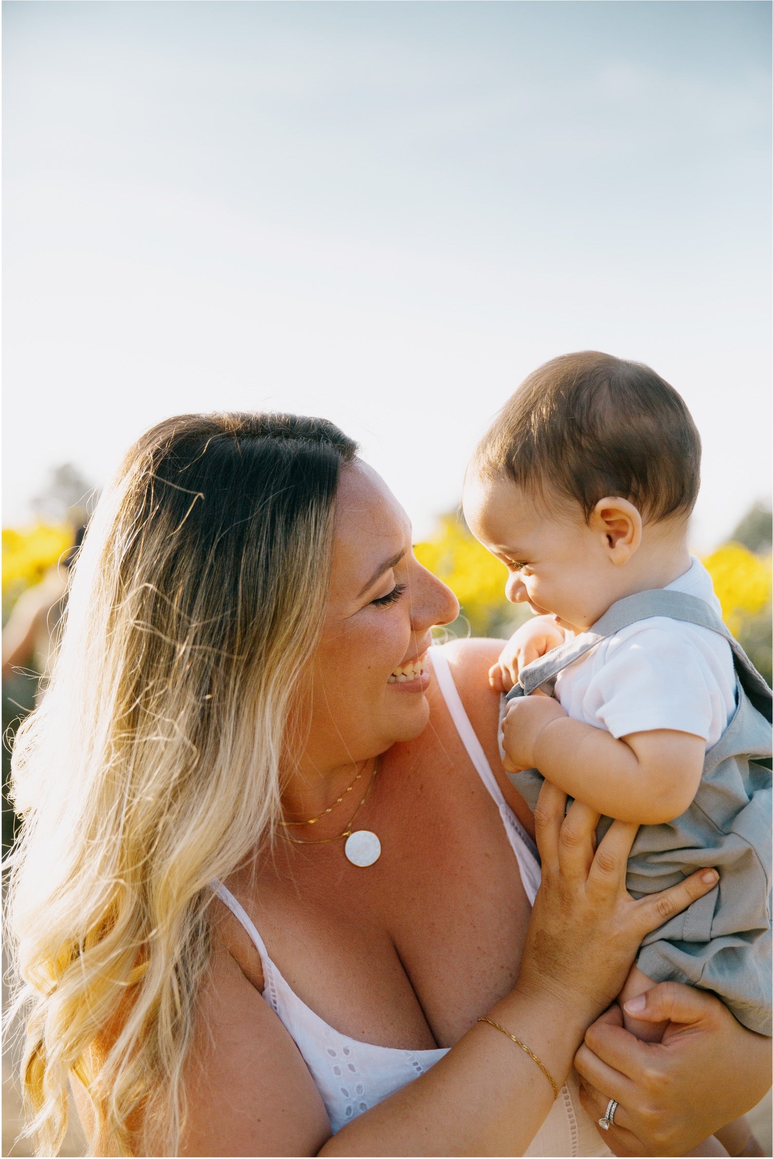 A smiling blonde woman outdoors holds a baby dressed in a white shirt and gray vest. The background is softly blurred with bright yellow flowers and a pale sky.