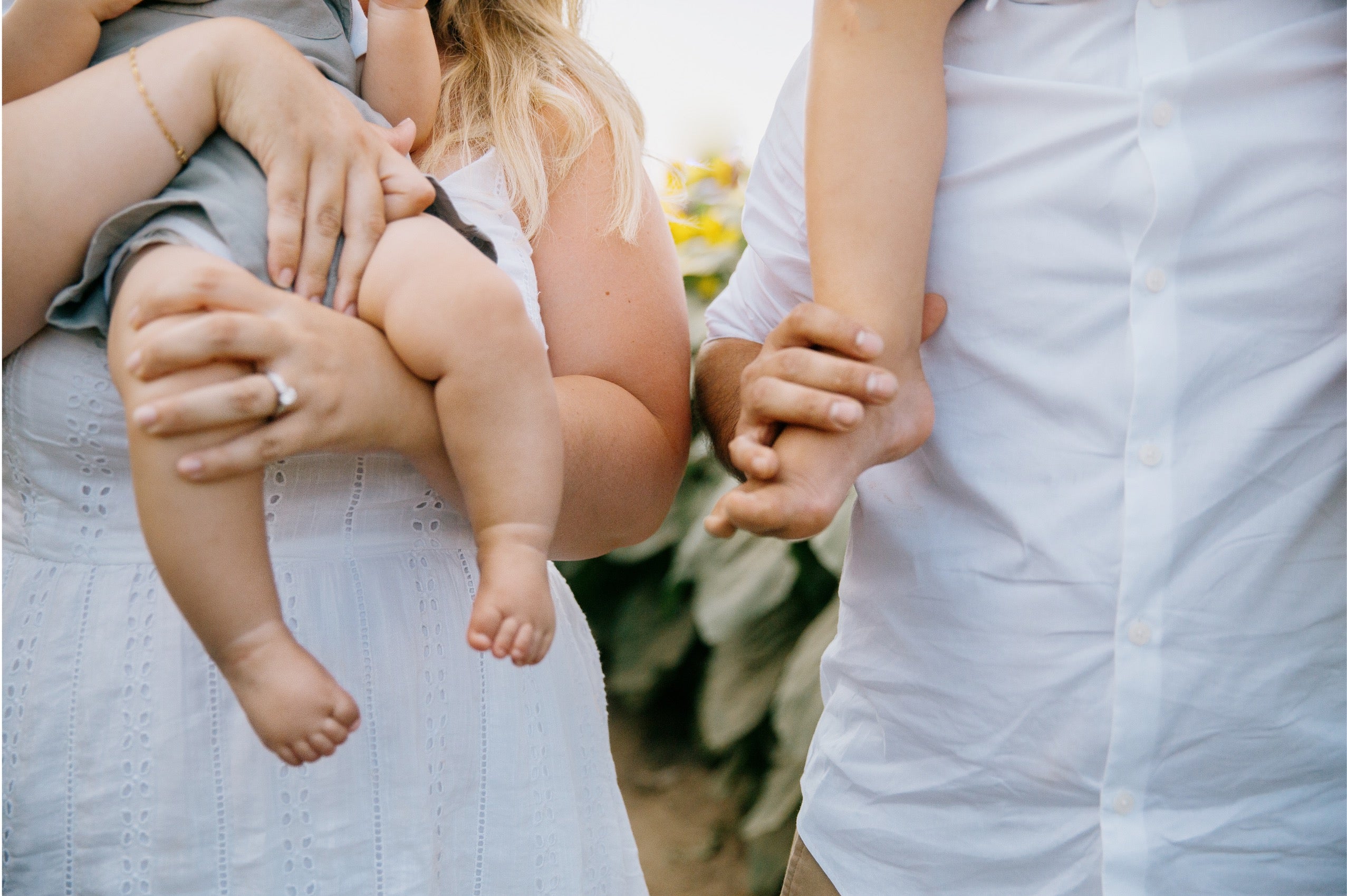 A close-up lifestyle shot of a family outdoors, featuring a woman in a white eyelet dress holding a baby, next to a man in a white button-down shirt holding a toddler.