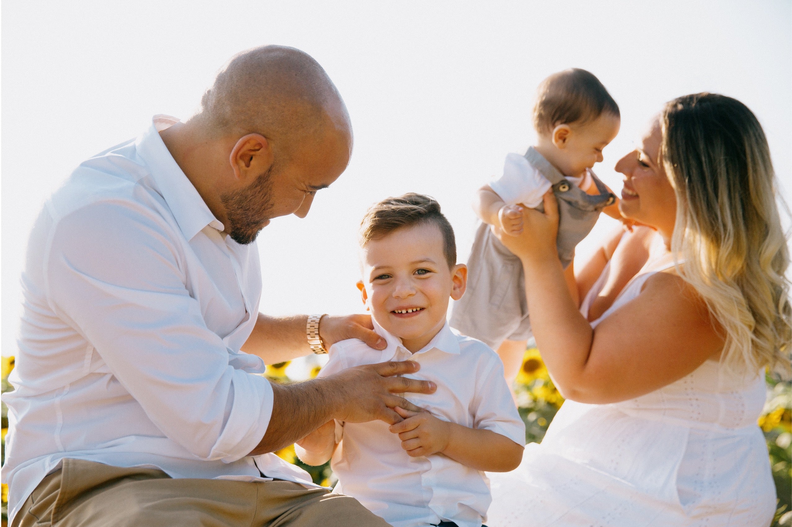 A family of four in white clothing posing outdoors in a sunflower field under bright sunlight. The father on the left is looking down at their son, who is smiling and wearing a white collared shirt.