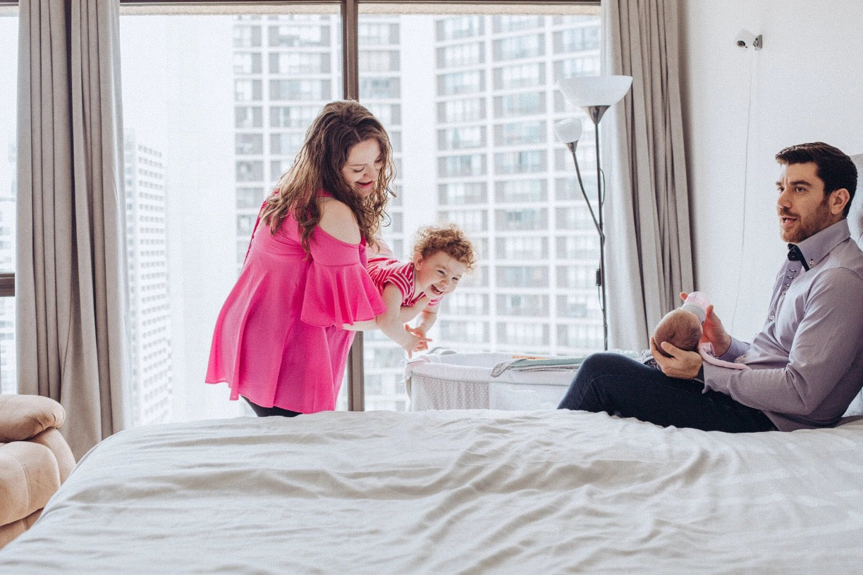 A family of three in a modern bedroom setting. A woman in a vibrant pink off-the-shoulder dress stands by the window holding a young child, while a man in a light purple shirt sits on the bed holding a baby.