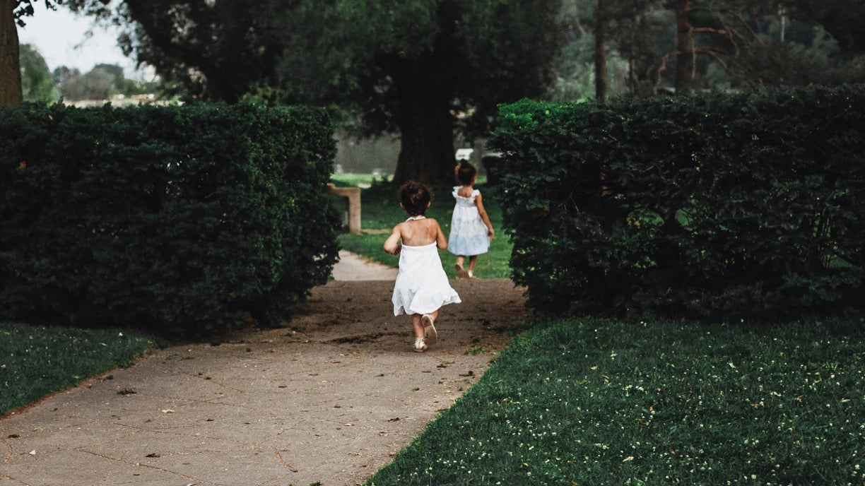 Two young girls in white dresses running down a dirt path between tall green hedges in a park setting.